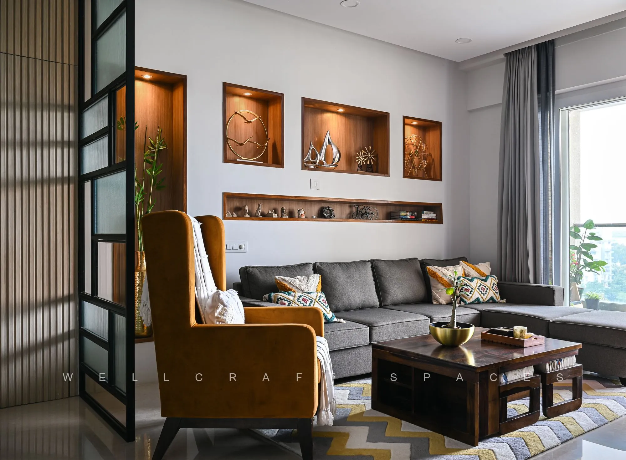 Living room with a grey sectional, tan wing chair, backlit walnut wall niches holding artefacts and a black-framed glazed partition.