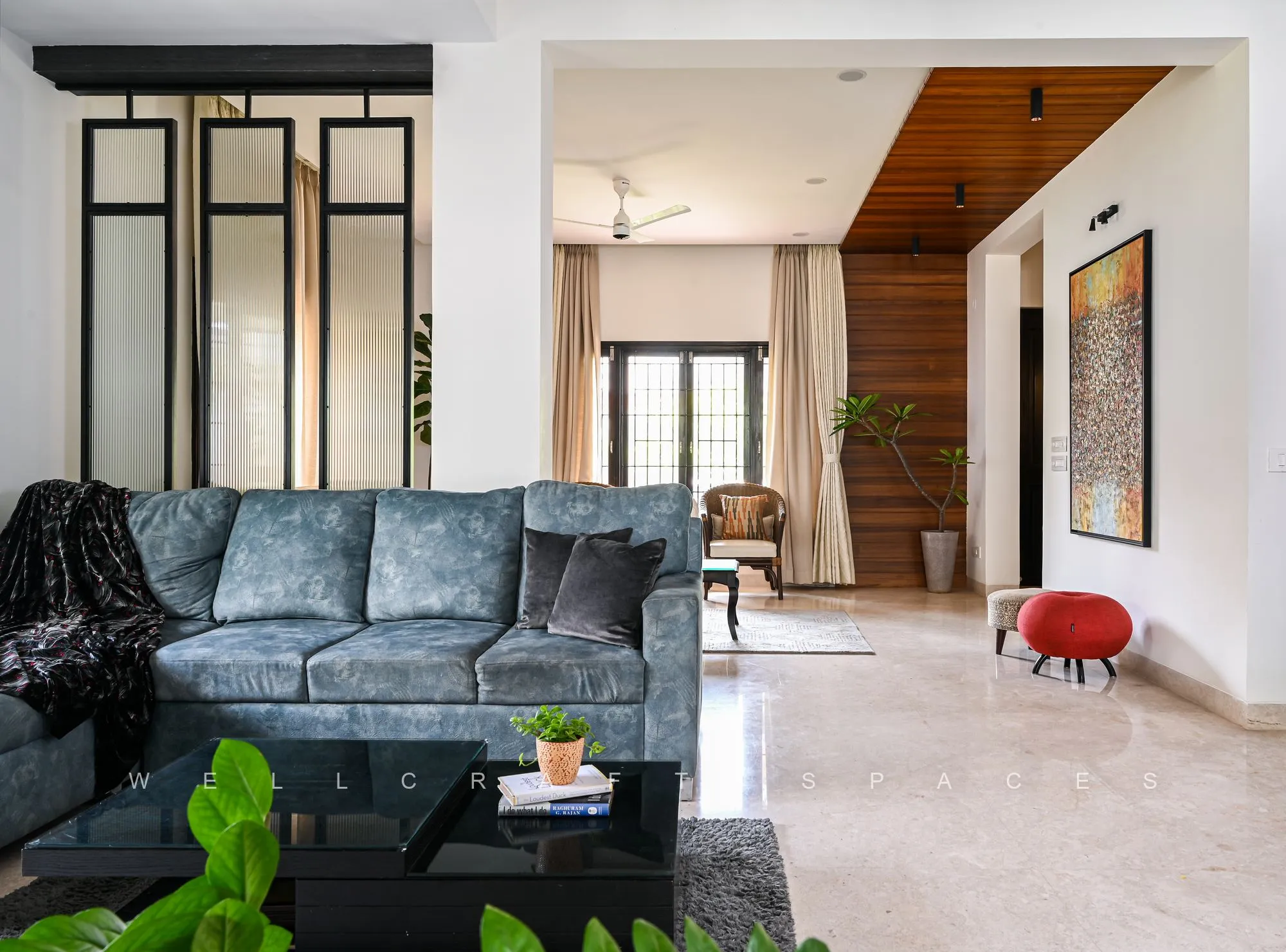Open living room in charcoal velvet sofas and cane armchairs, with a black-framed glass partition and a timber-clad staircase wall drawing the eye deeper into the house.