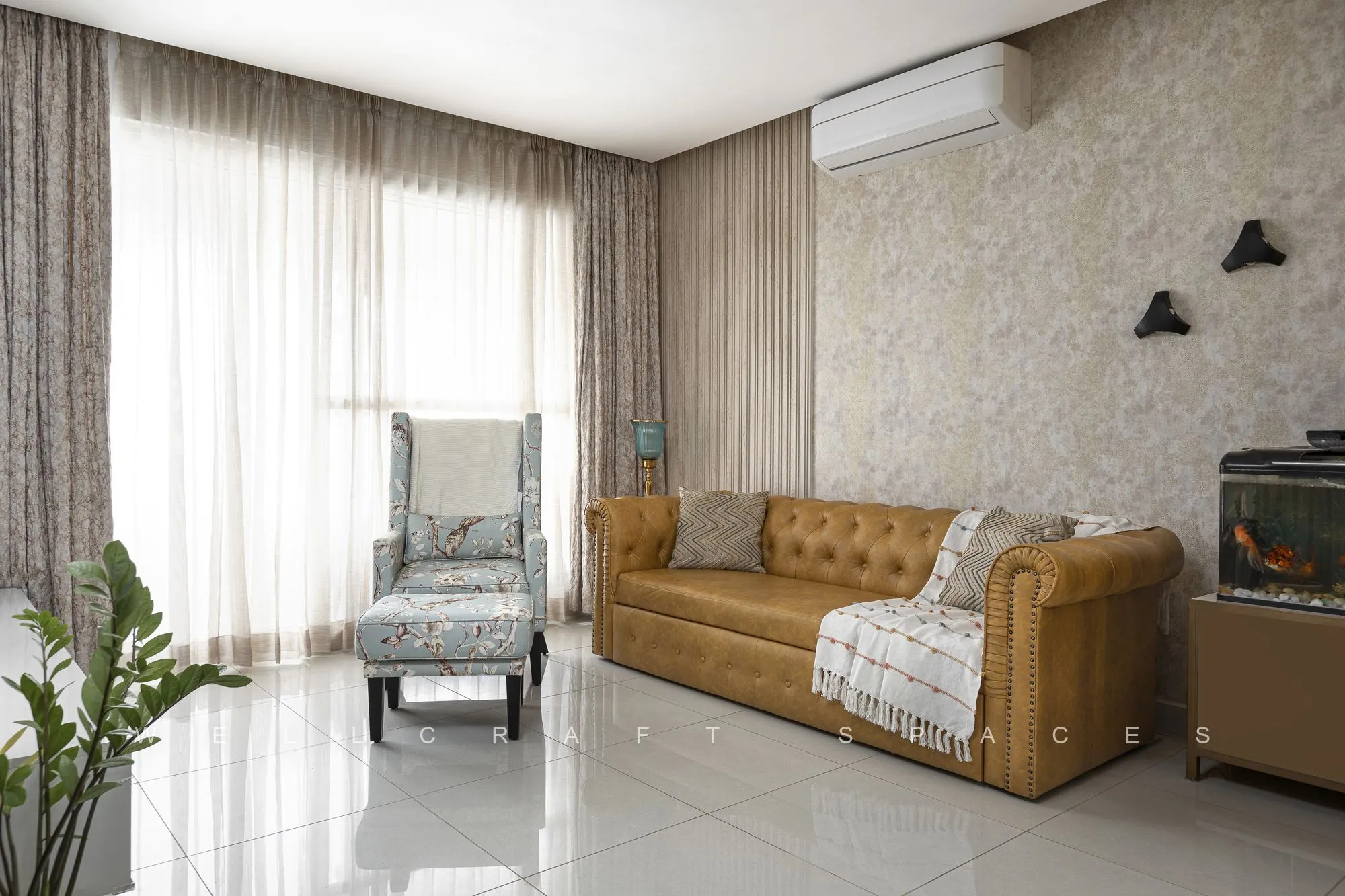 Bright living room with a tan leather chesterfield sofa, a grey wingback chair and sheer drapes over glossy ivory floor tiles.