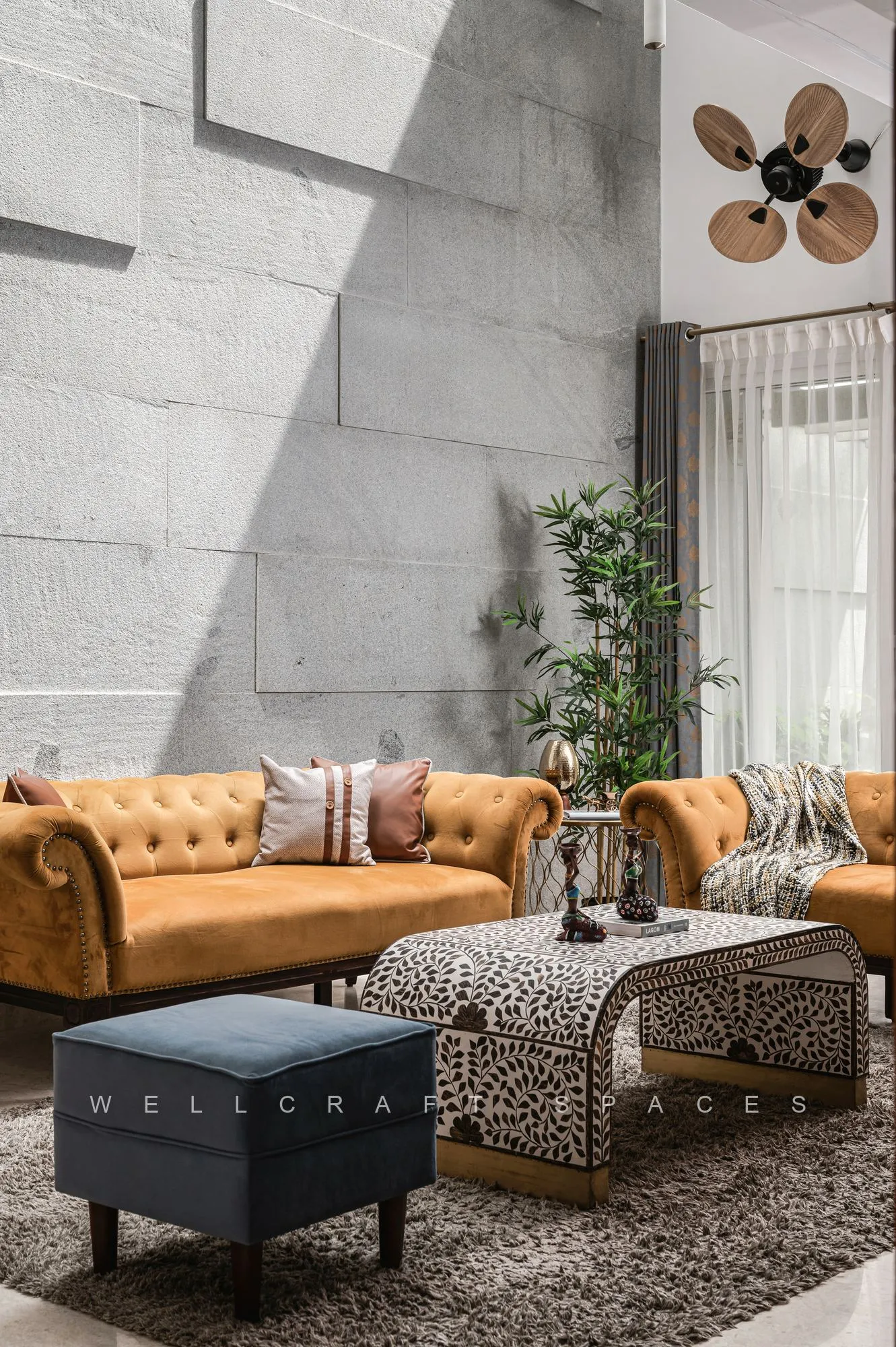 Mustard velvet tufted sofa and a navy ottoman set against a full-height board-formed concrete wall, with animal-print stools, a brass sunburst mirror and tall potted greenery.