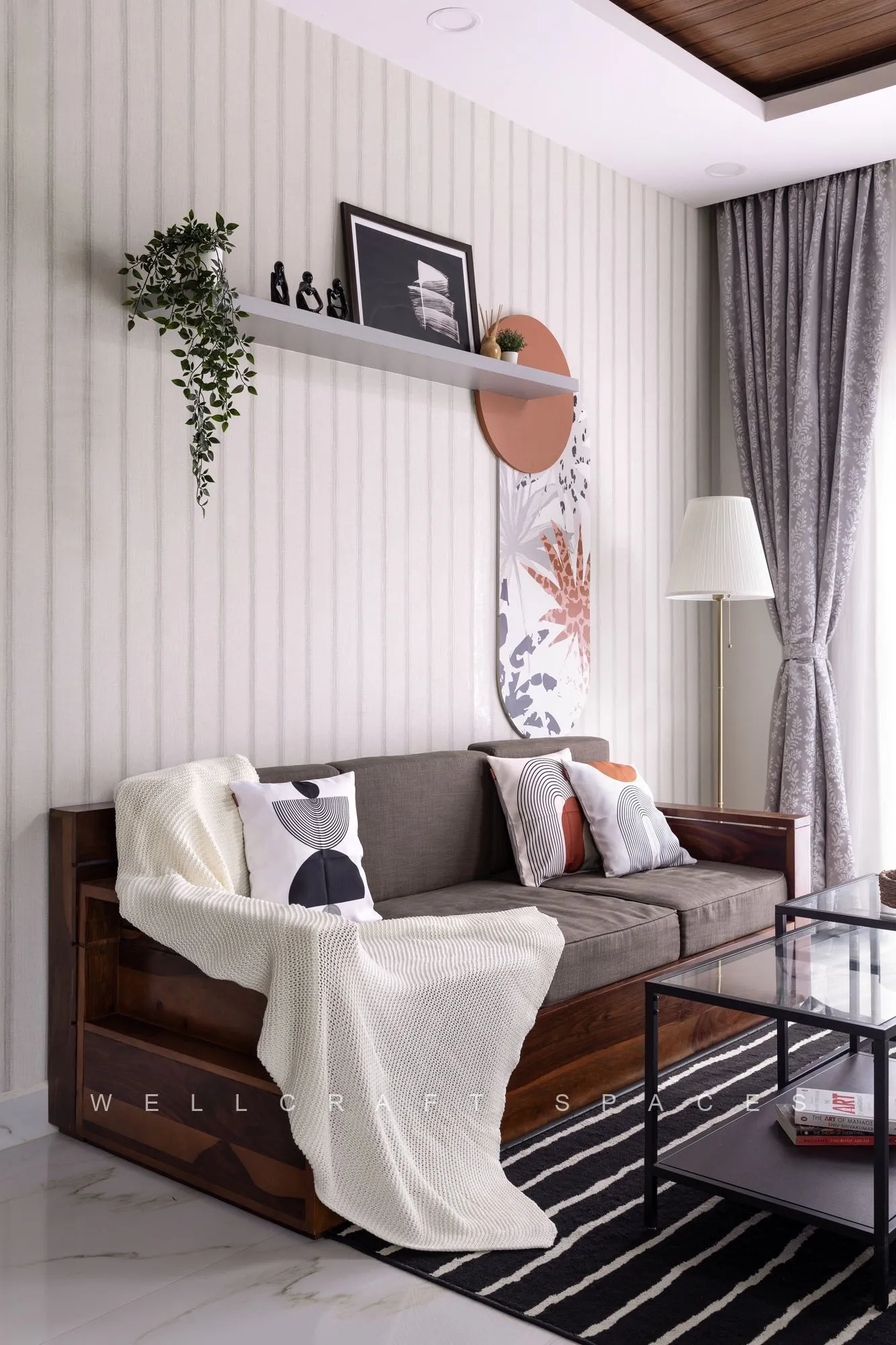 Living room with a teak-framed sofa in taupe upholstery, a cream throw and a black-and-white striped rug, set against fluted white panelling and sheer grey drapes.