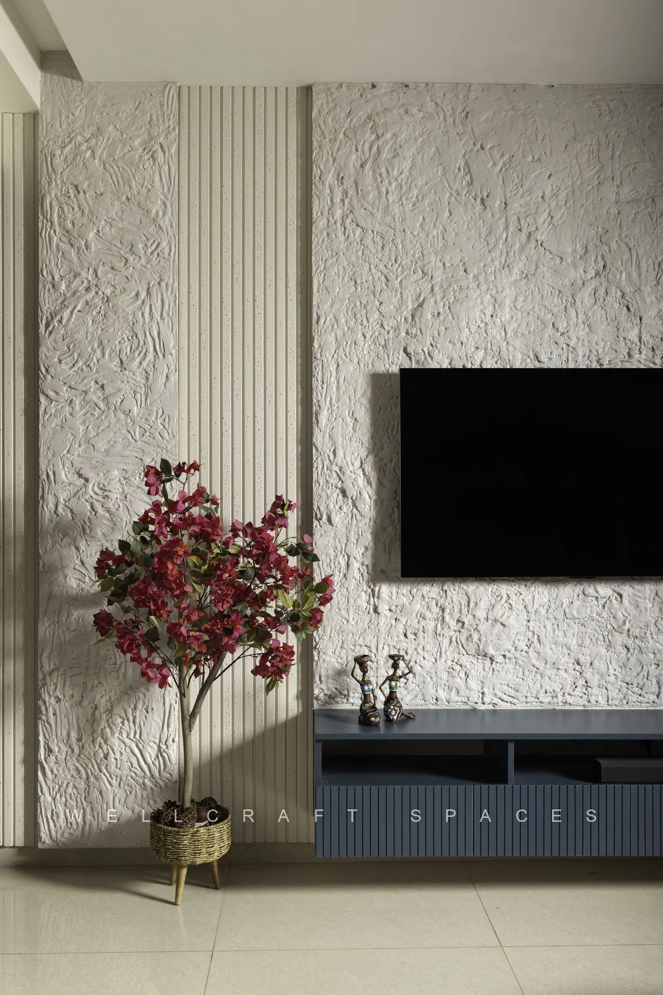 Television wall in rough white textured plaster with a fluted side panel, a deep blue floating console and a tall red blossom tree in a brass planter.