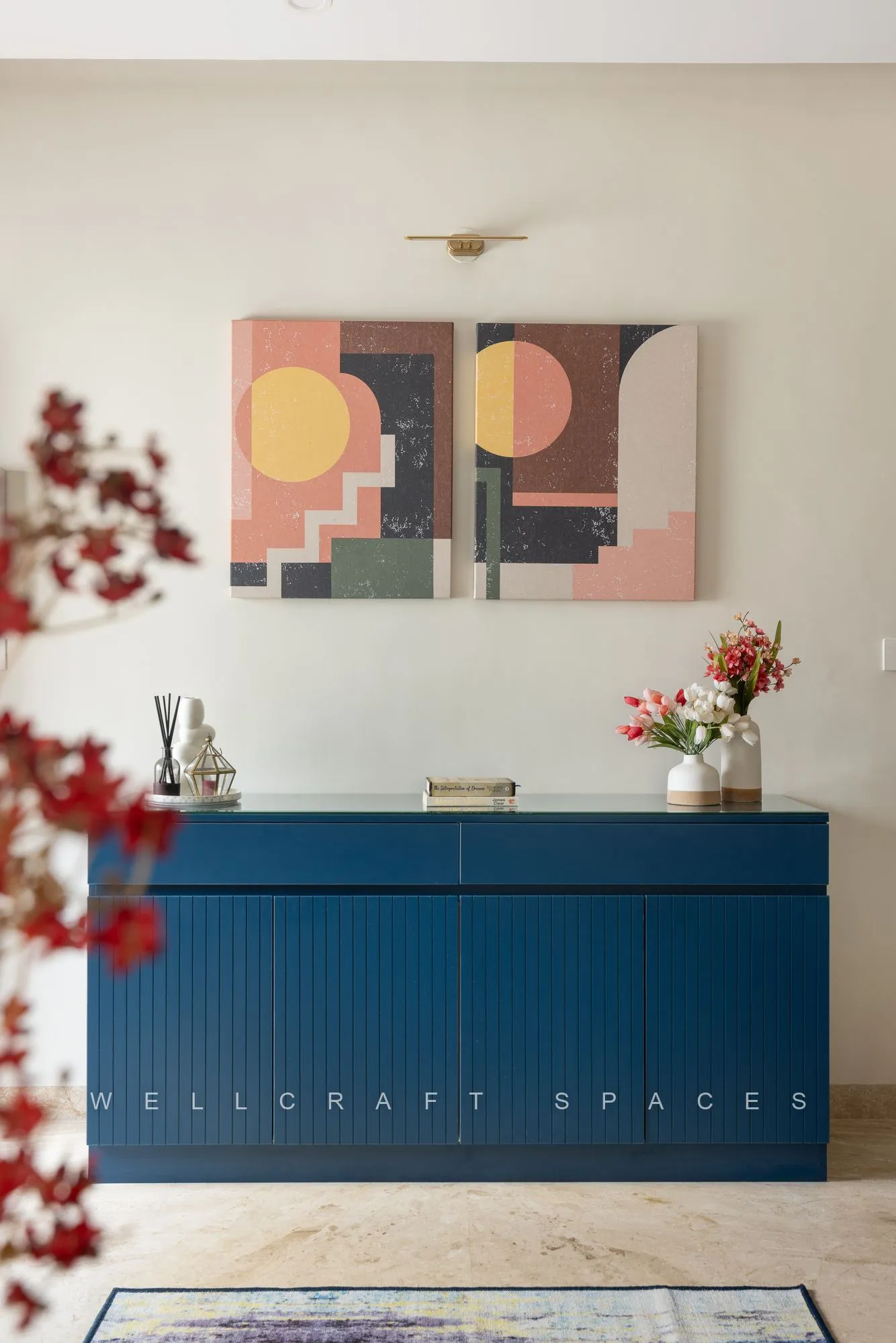 Fluted console in deep petrol blue against a white wall hung with two muted geometric canvases, dressed with fresh blossom and a marble-look floor.