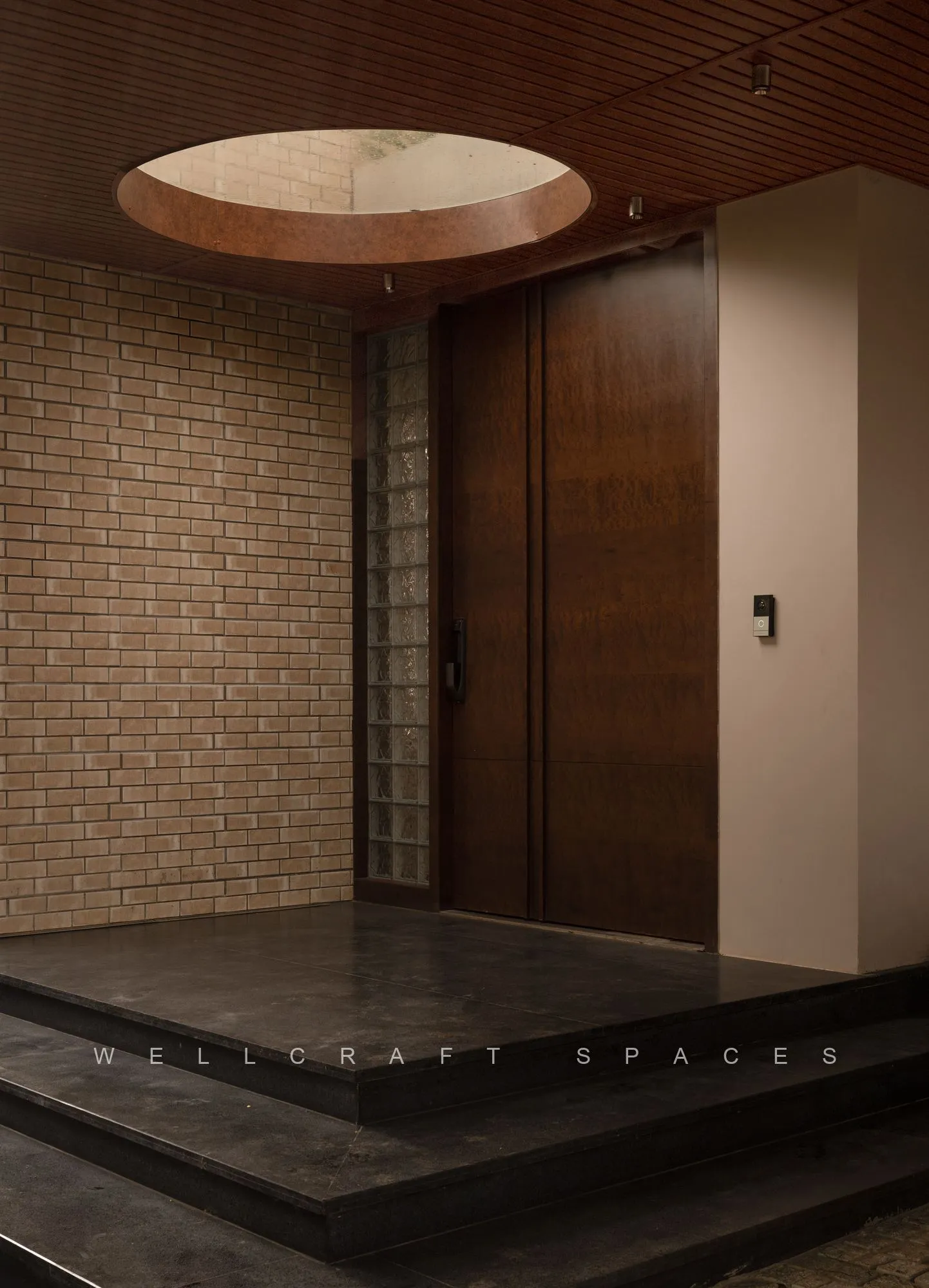 Entrance porch clad in narrow brick, with a tall solid teak door, dark stone steps and a circular wooden ceiling medallion above.