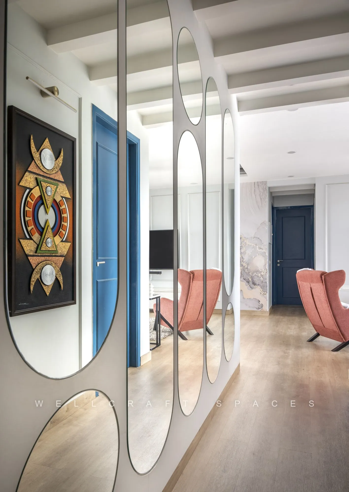Entrance passage lined with organic-shaped mirrors and a jewel-toned mandala artwork, opening onto the living room and its blush velvet armchairs.