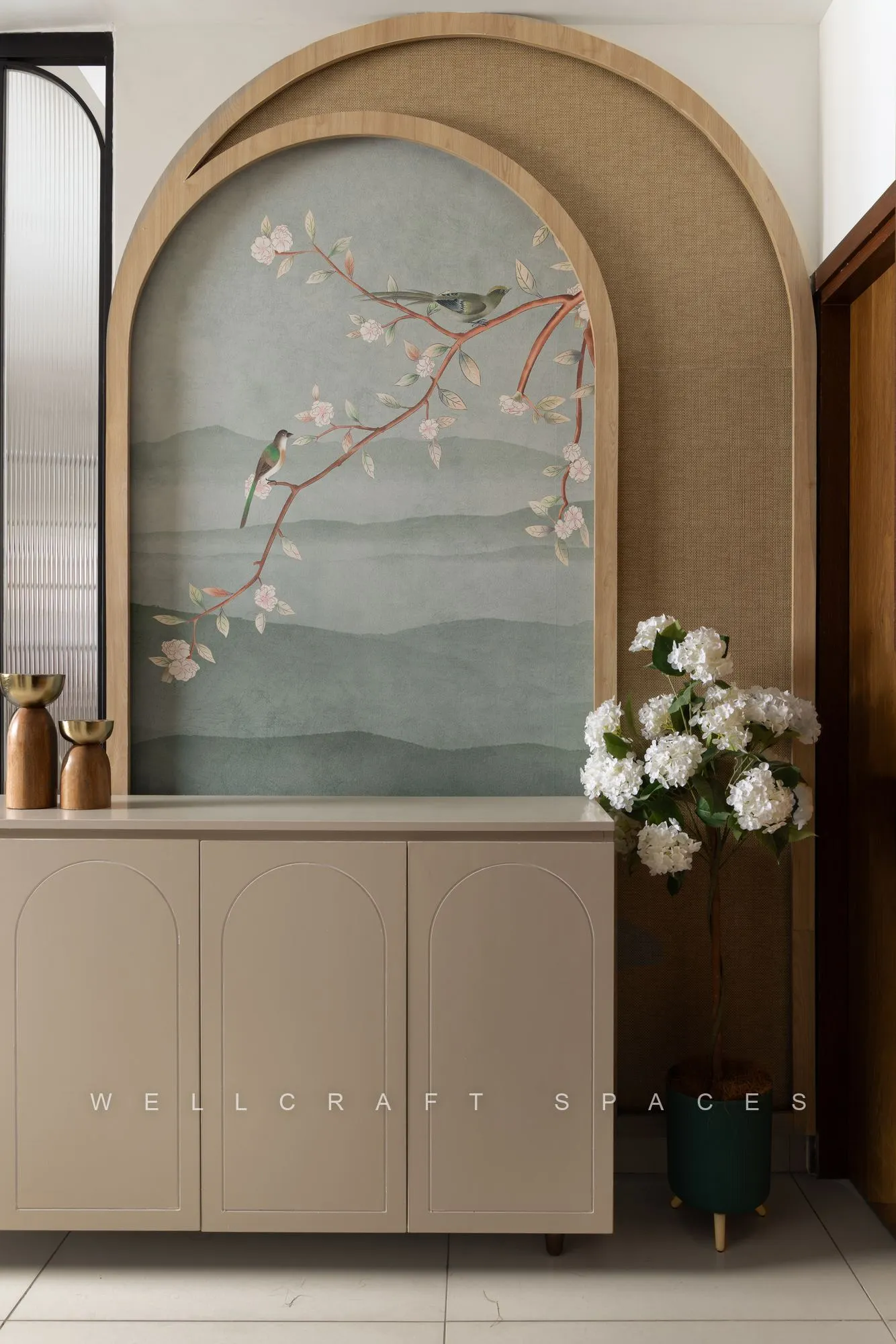 Entry vignette with an arched cherry-blossom mural panel framed in cane, a cream console and brass vases beside white blooms.