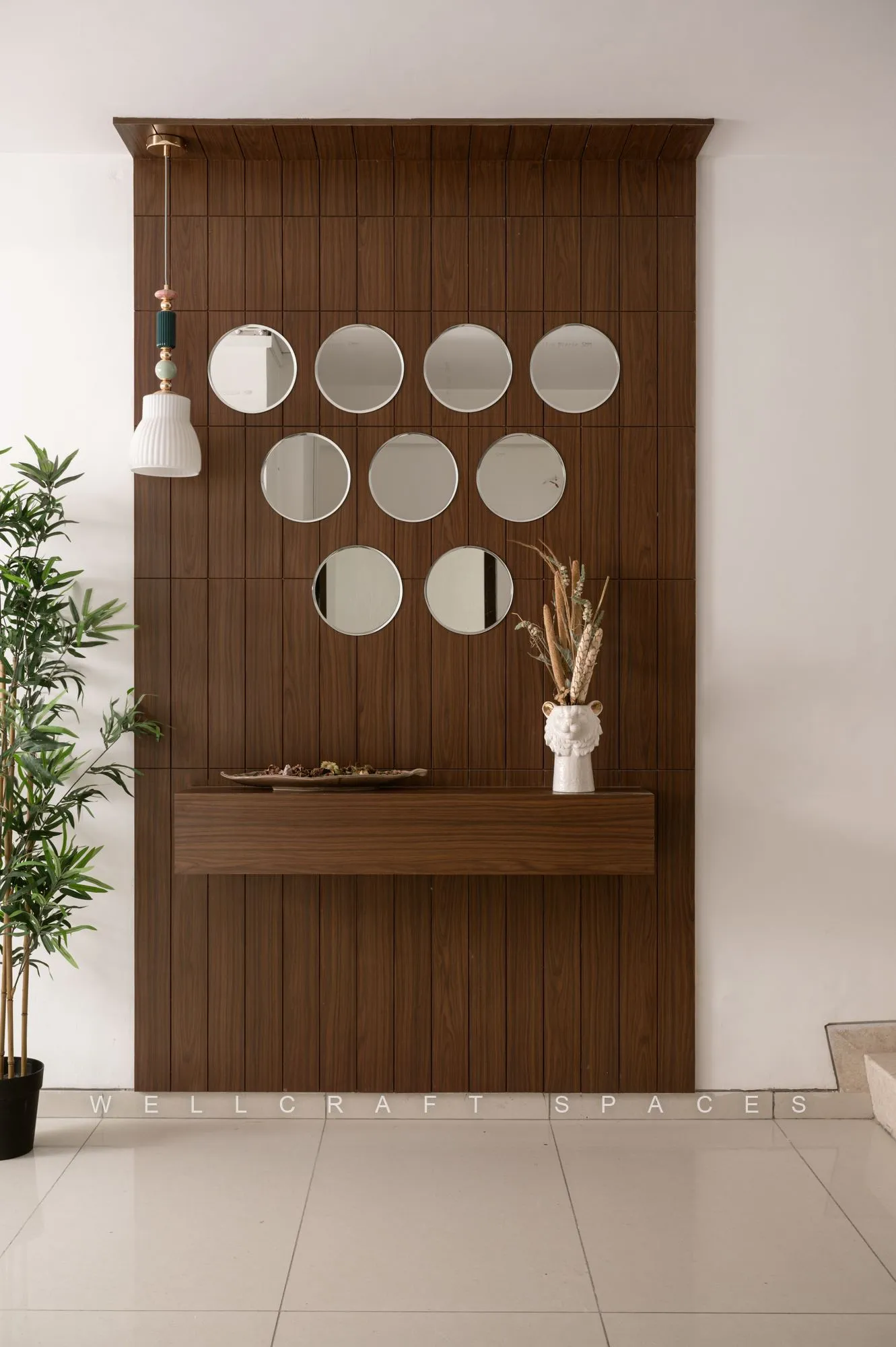 Basement foyer with a vertically slatted walnut screen hung with a cluster of round mirrors, a white ceramic vase of dried stems and glossy ivory floor tiles.