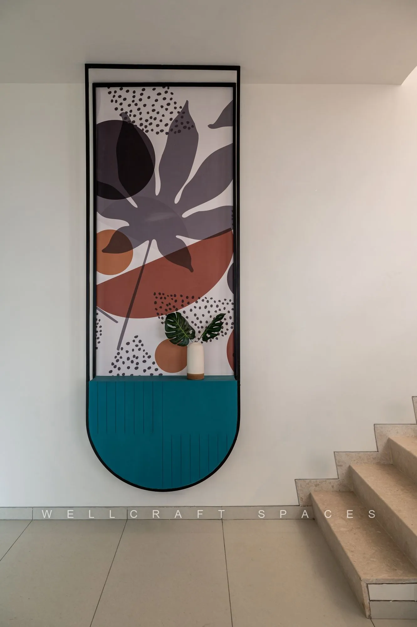 Entrance landing beside the timber staircase, with a tall abstract panel in plum and teal above a planter, against plain white walls and ivory floor tiles.
