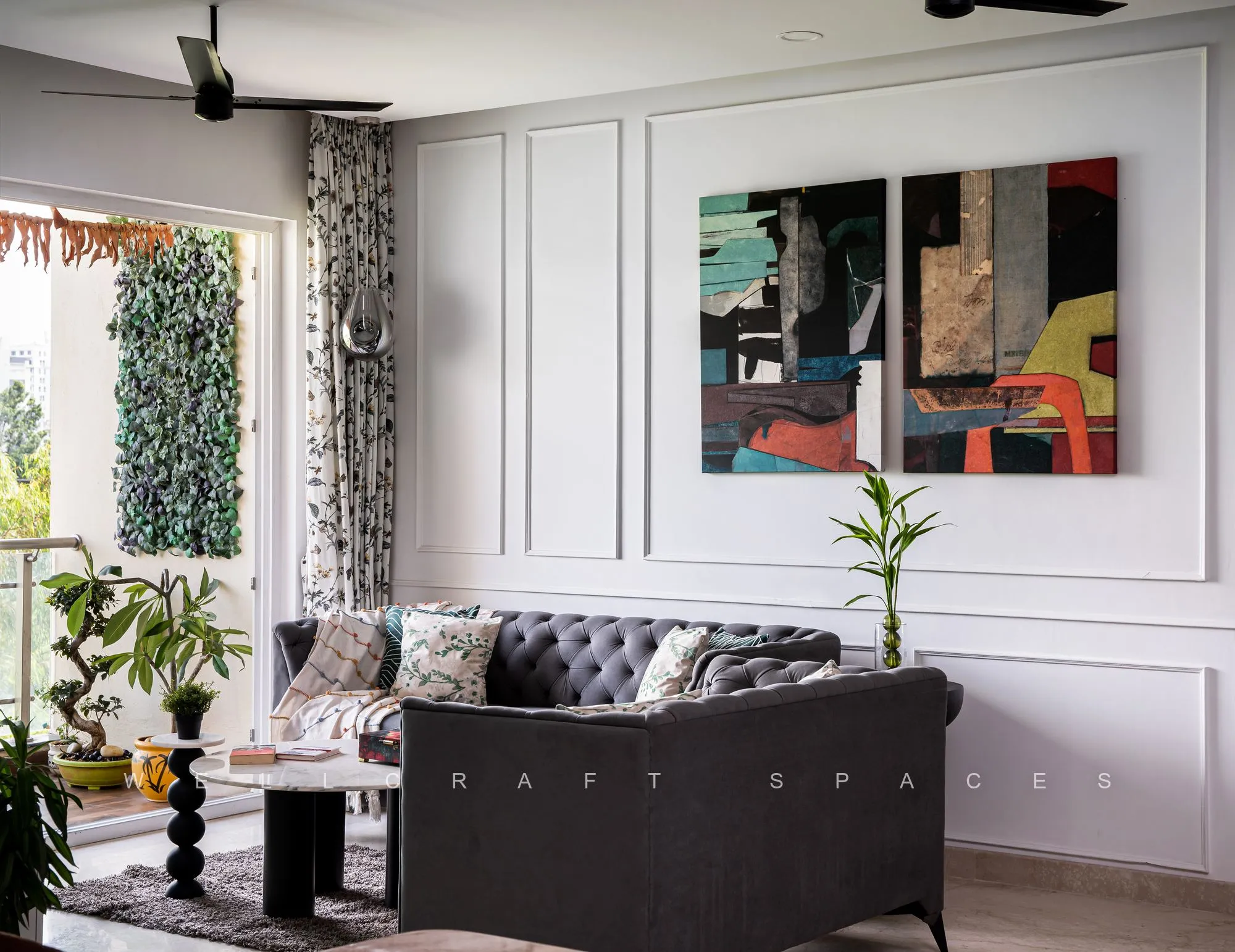 Charcoal velvet chesterfield sofa with printed cushions and a black marble side table, set against white panelled walls hung with a colourful abstract canvas.