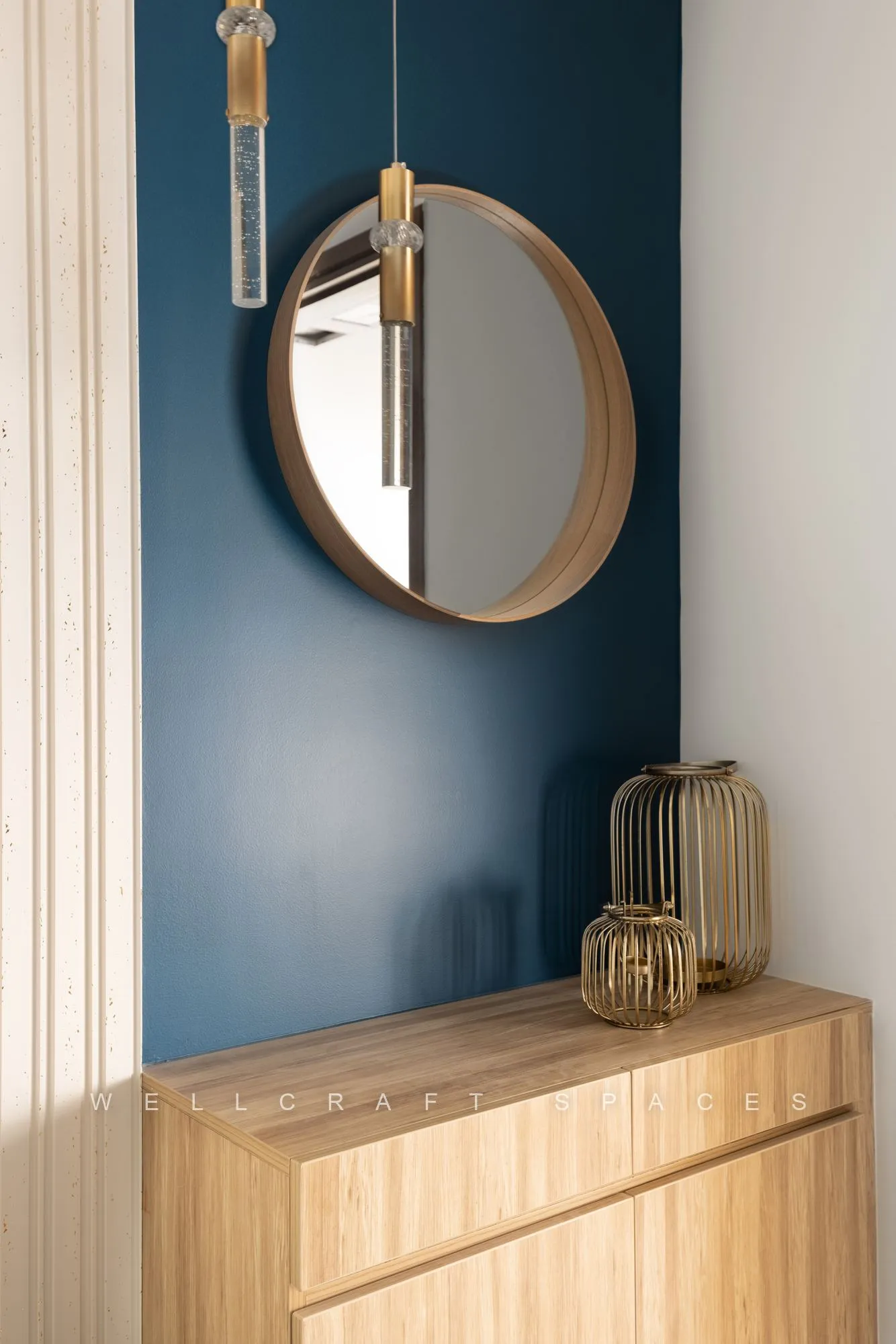 Entry console in light oak against a deep petrol-blue wall, topped by a round brass-framed mirror and a slender glass pendant.
