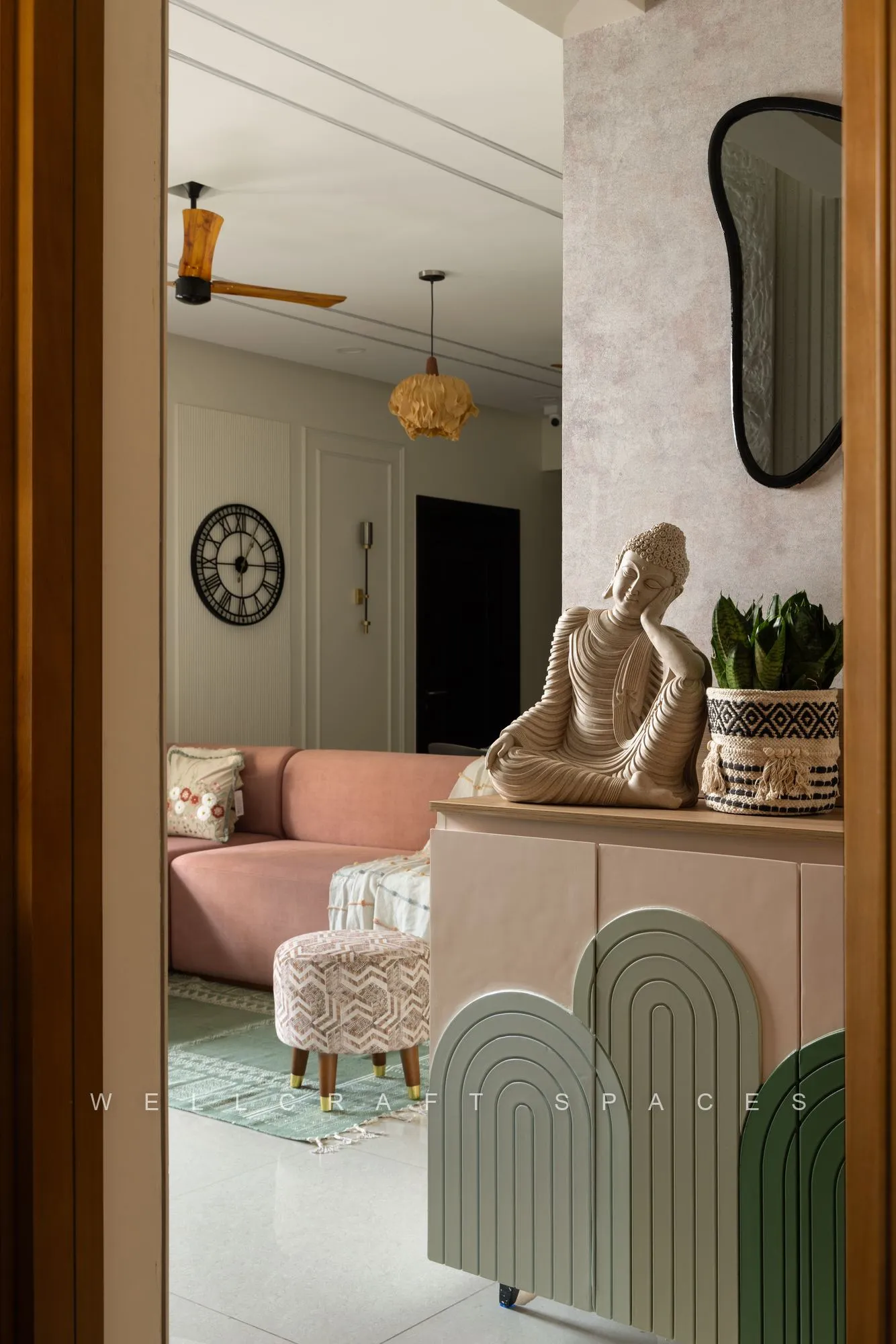 Entry framed by a teak door, with a seated stone figure on an arched sage-and-cream console unit, a black round mirror and a pink sofa beyond.