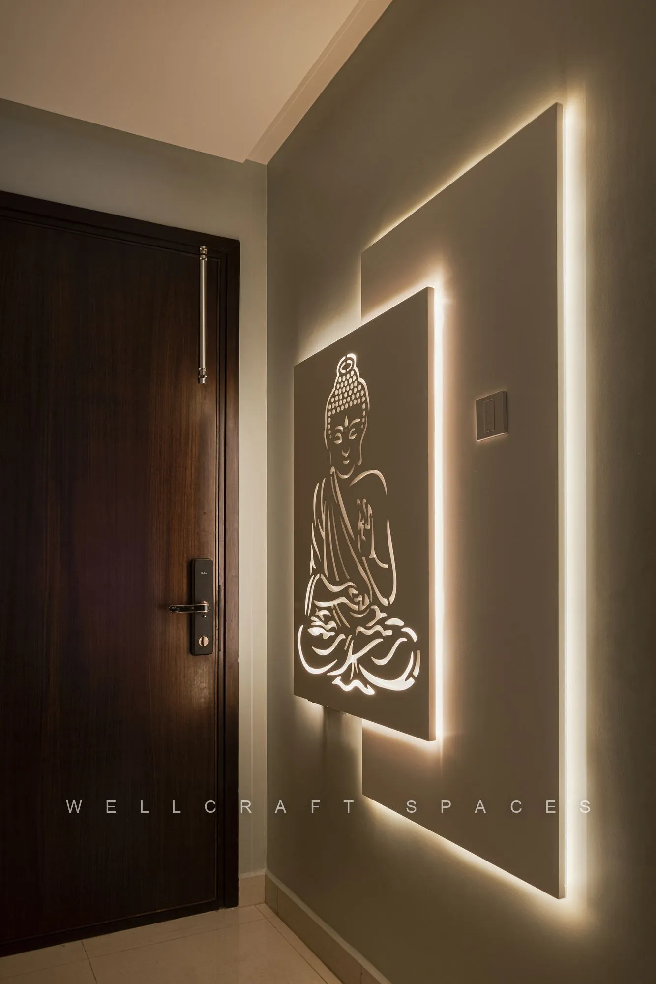 Dark teak entrance door beside a backlit wall panel with an etched meditating Buddha outlined in warm cove lighting.
