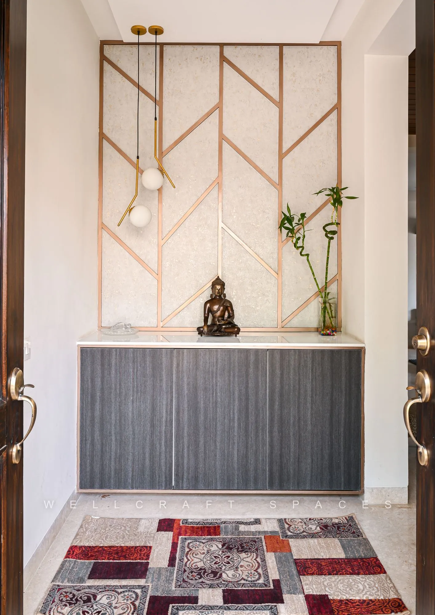 Entrance console in dark ribbed wood beneath a geometric gold-inlay wall panel, with a brass Buddha, pendant bulbs and a red patterned runner on ivory tile.