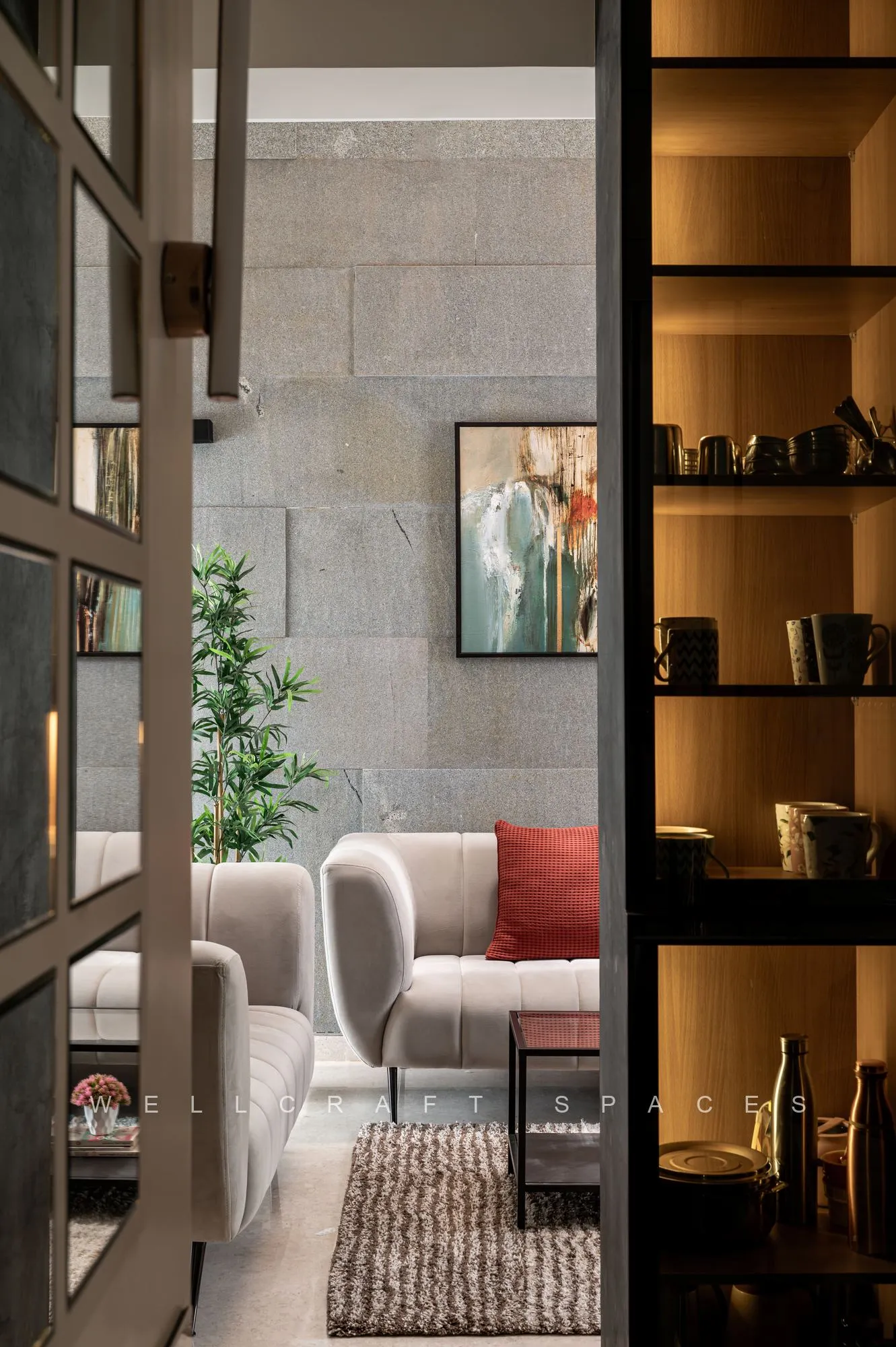 View into the living room through a black metal and glass screen, past a backlit display shelf holding dark ceramics, to the cream sofa and shaggy grey rug.