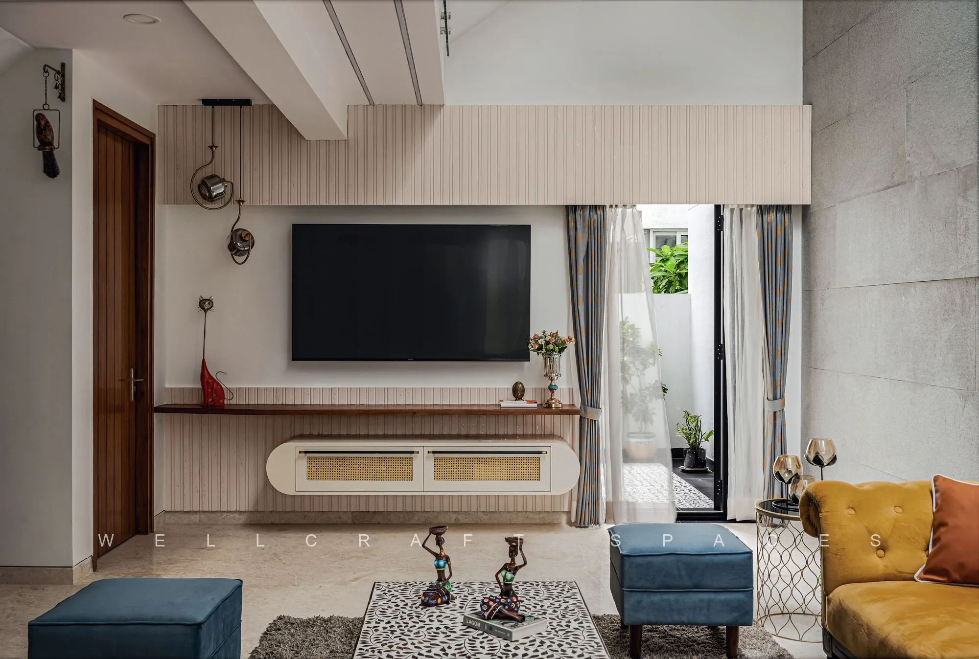 Wide view of the living room showing the panelled media wall with a floating ledge, low blue and printed ottomans and sliding glass doors to the garden.
