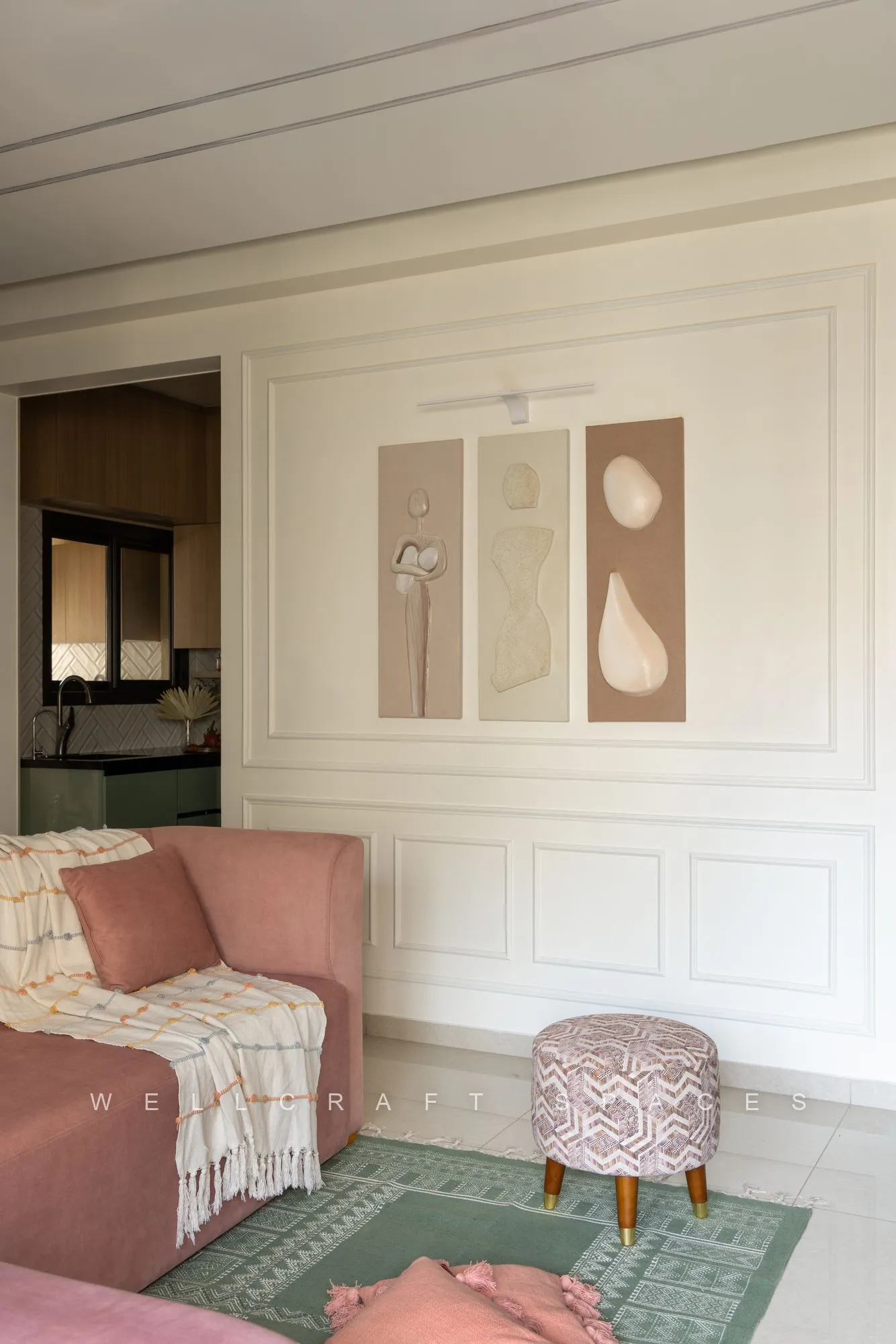 Living corner with a dusty pink sofa, a white moulded panel wall hung with abstract prints and a striped ottoman on a green rug.