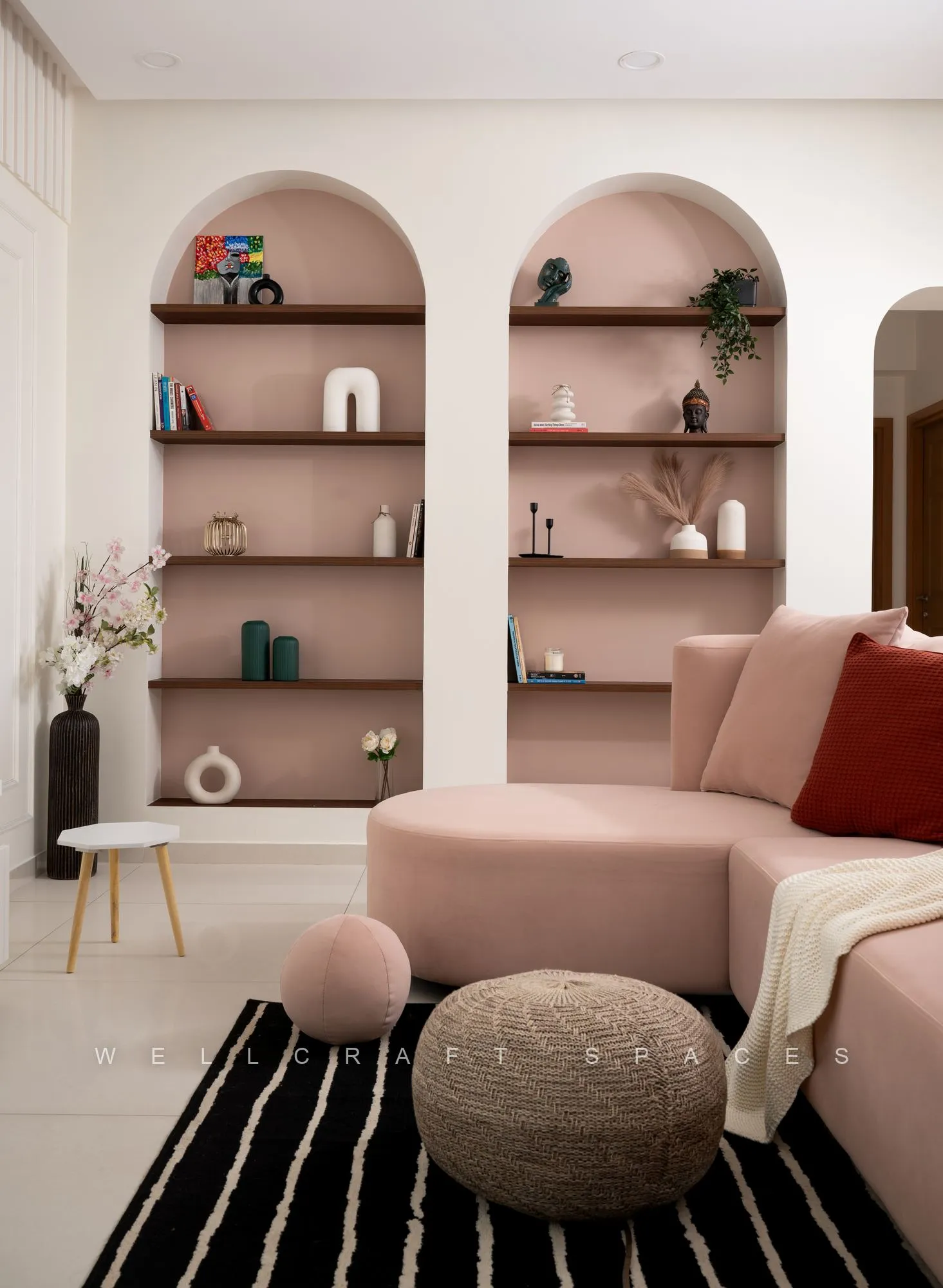 Curved pink sectional with a red cushion and knitted pouffe, set against twin arched niches styled with ceramics and greenery.