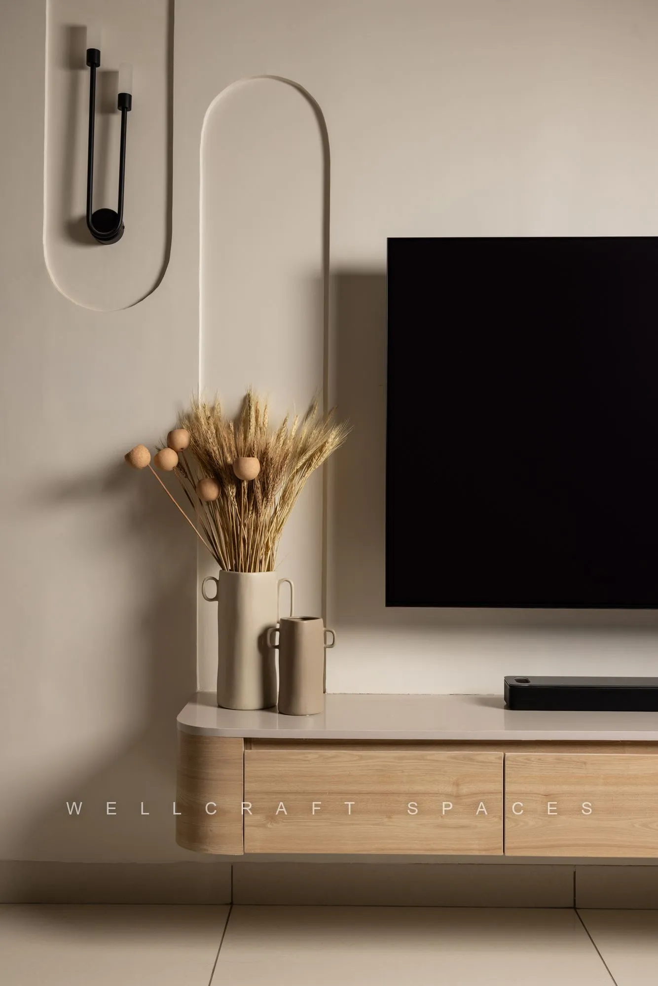 Television mounted within a rounded cream wall profile above a floating oak ledge, styled with dried grasses and a slim black sconce.