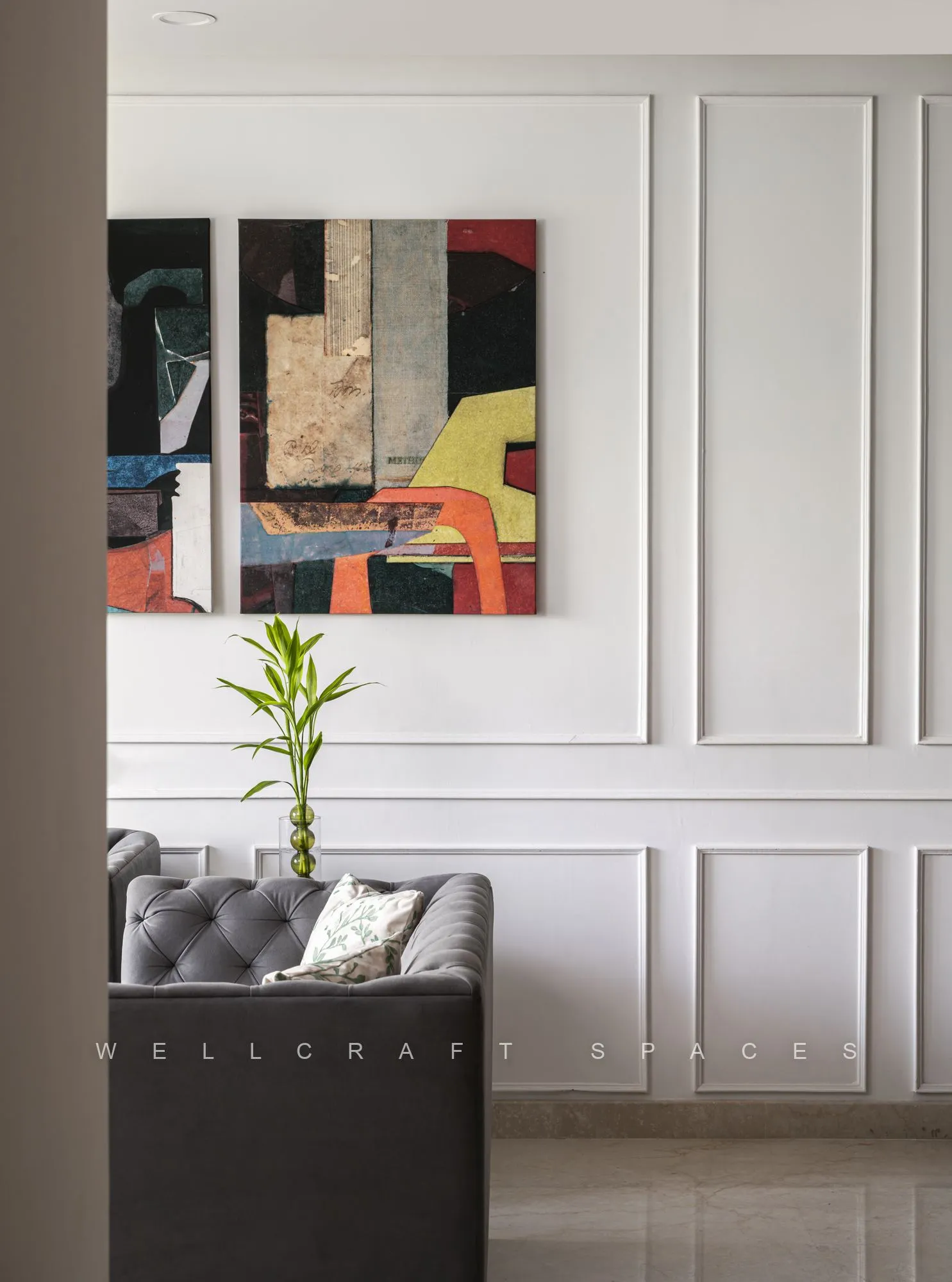 Corner of the living room where the grey tufted sofa meets a white panelled wall carrying two abstract artworks, with a slim potted dracaena between them.