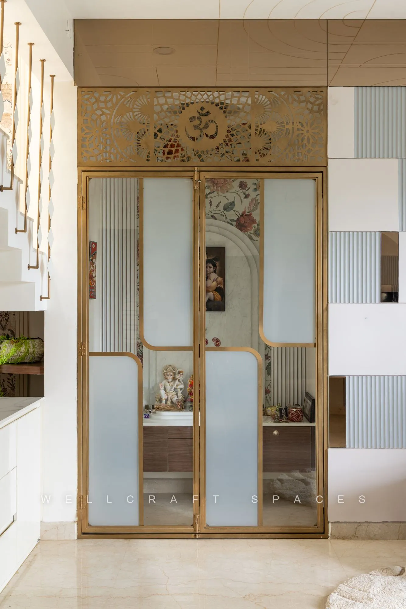 Pooja unit set in a mirrored and fluted glass surround with a brass frame, gold floral wallpaper above and a walnut counter holding the idols.