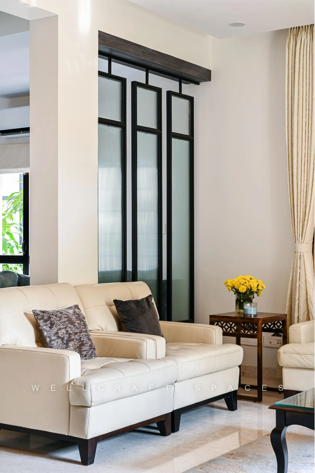 Formal seating corner with a cream two-seater, patterned cushions and a marble-topped side table, set against black-framed windows and full-height sheer drapes.
