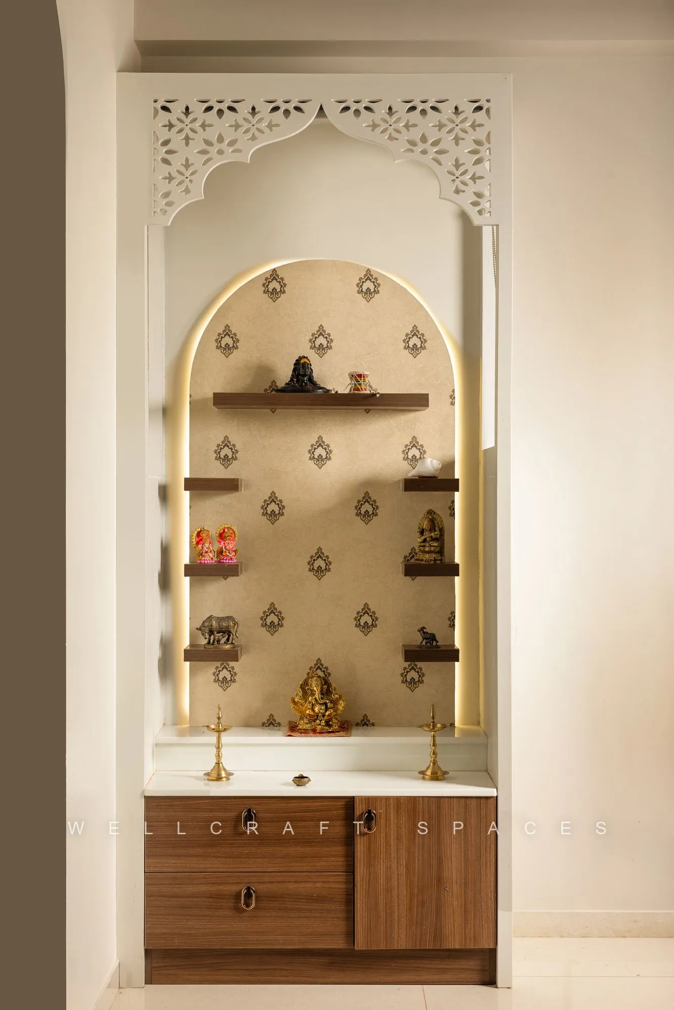 Arched pooja niche with carved white jaali surround, patterned gold backdrop and a walnut cabinet base holding brass idols.