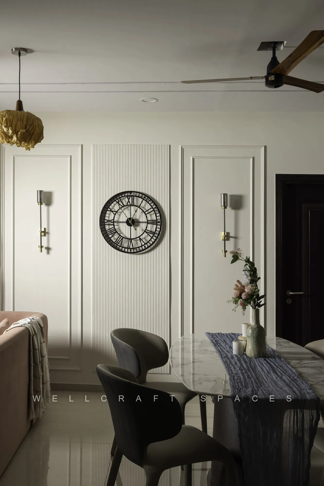Dining area with sculptural black chairs and a stone-top table against classic white wall mouldings, a round clock and brass wall lights.