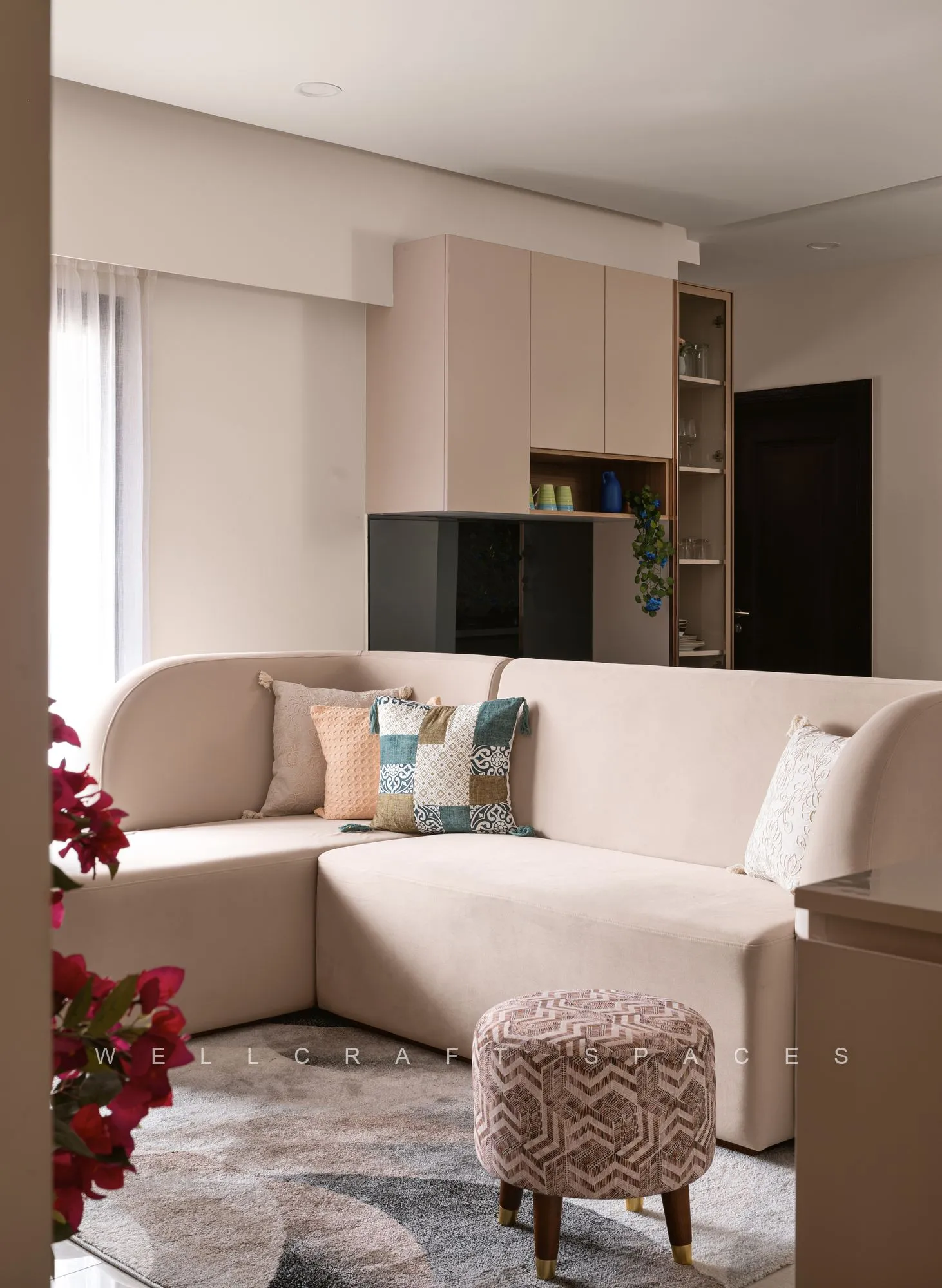 L-shaped cream sofa with printed cushions and a patterned rug, opening to a built-in media wall and a bright window beyond.
