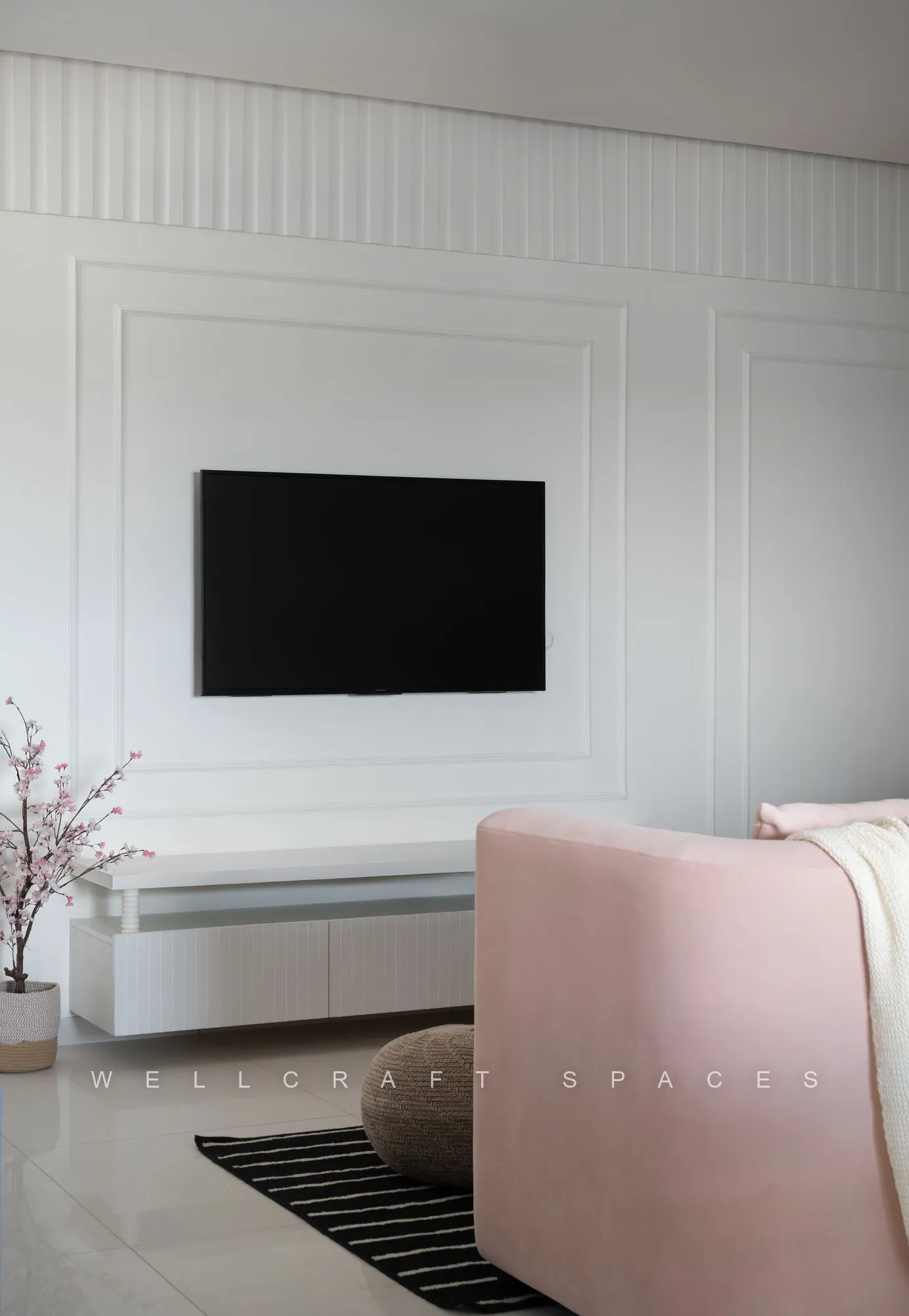 Television mounted on a white moulded panel wall above a floating console, with the edge of the blush sofa and striped rug in view.