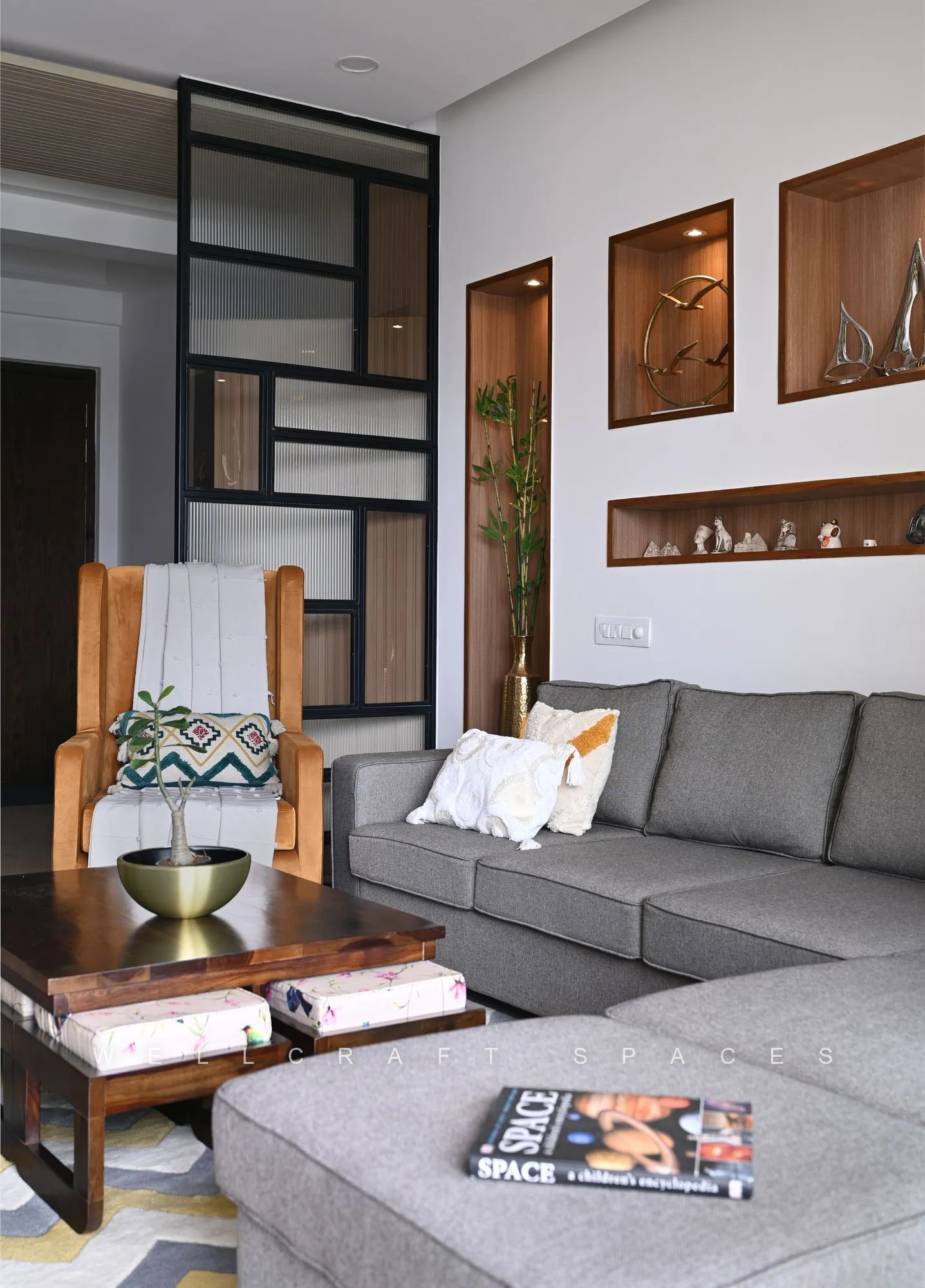 Grey L-shaped sofa with patterned cushions and a low walnut coffee table, set below walnut display niches and a black metal shelf frame.