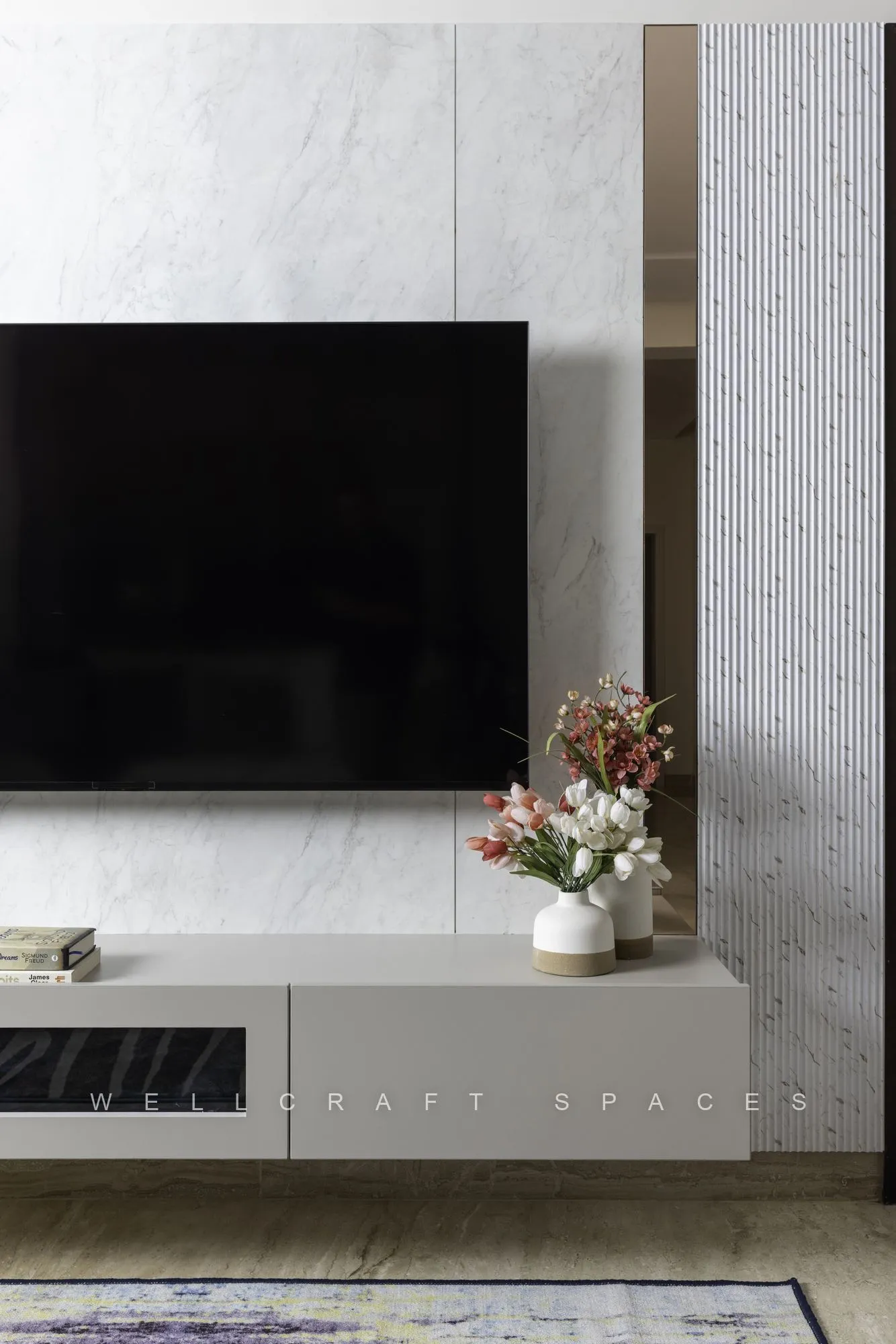 Media wall in book-matched white marble with a wall-hung television and a slim grey floating console holding a vase of blossom.