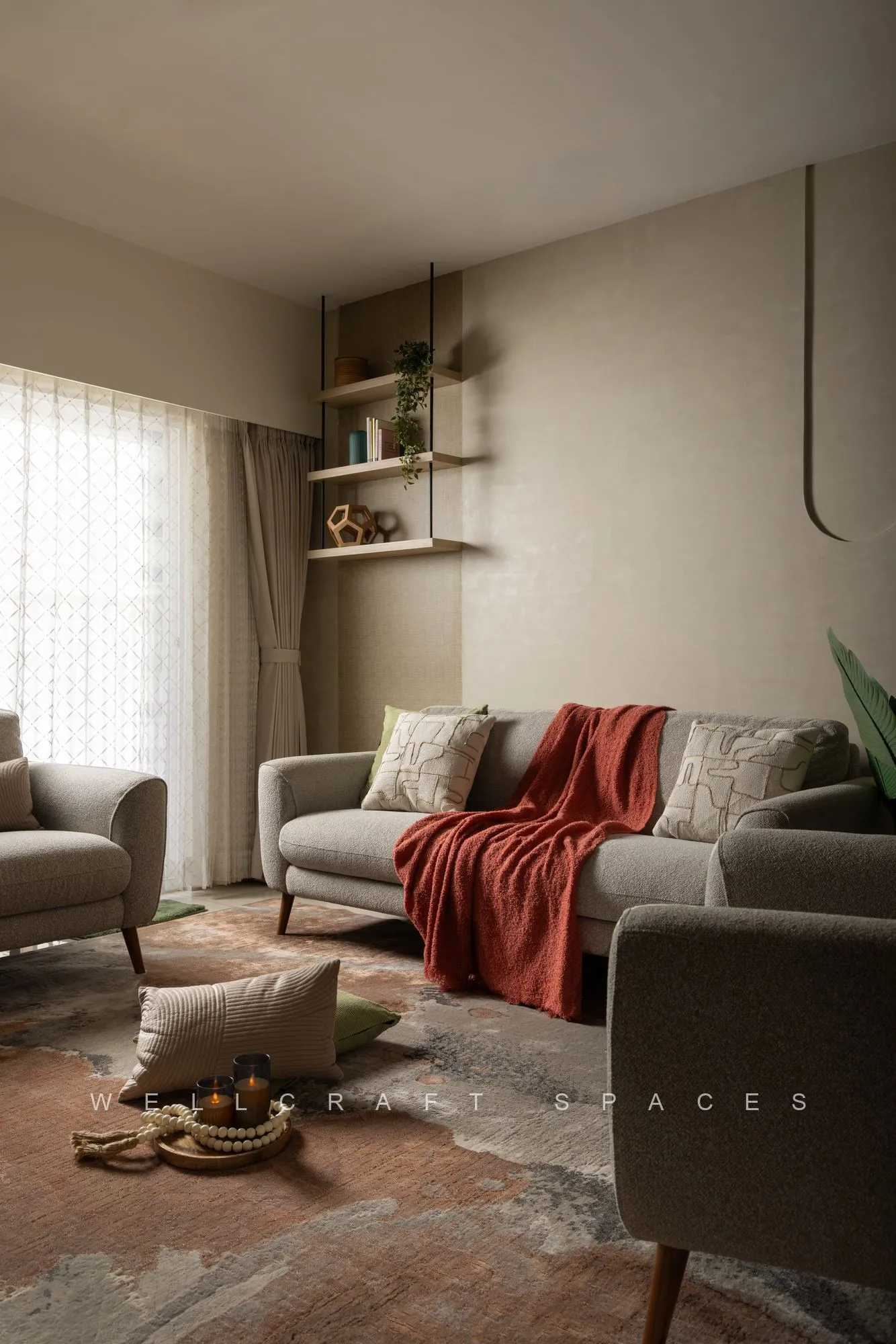 Grey sofa layered with a rust throw and cushions on a faded pink rug, with open wall shelves and a curtained window behind.