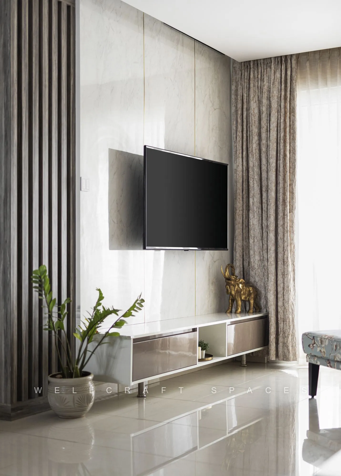 Television on a textured panelled wall above a slim floating console, with potted greenery and full-length taupe curtains.