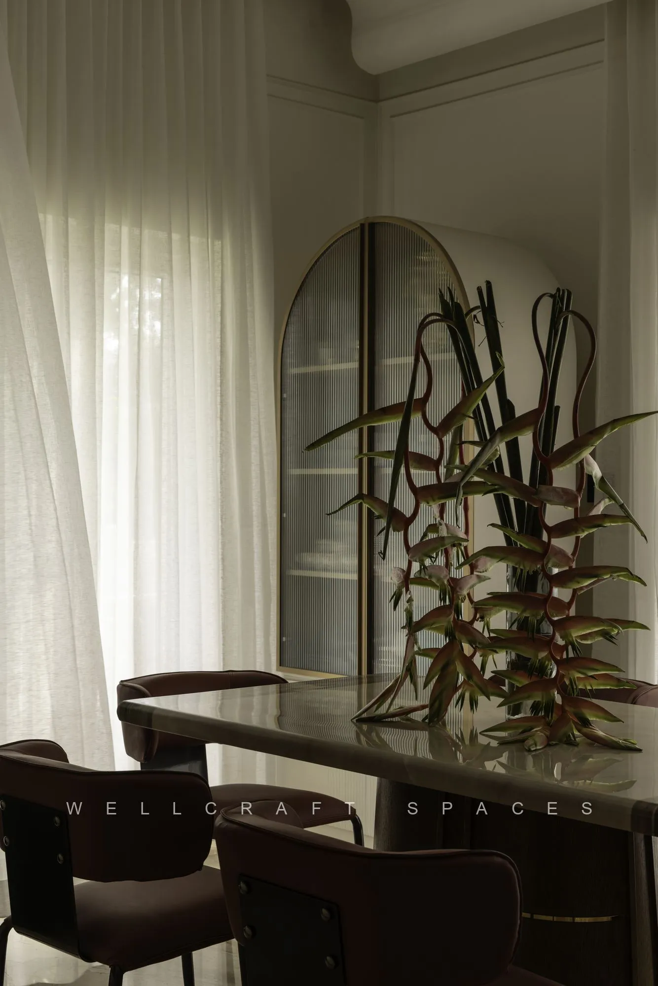 Dining table in dark stone with brown leather chairs, a tall sculptural branch centrepiece and a black arched mirror against sheer curtains.