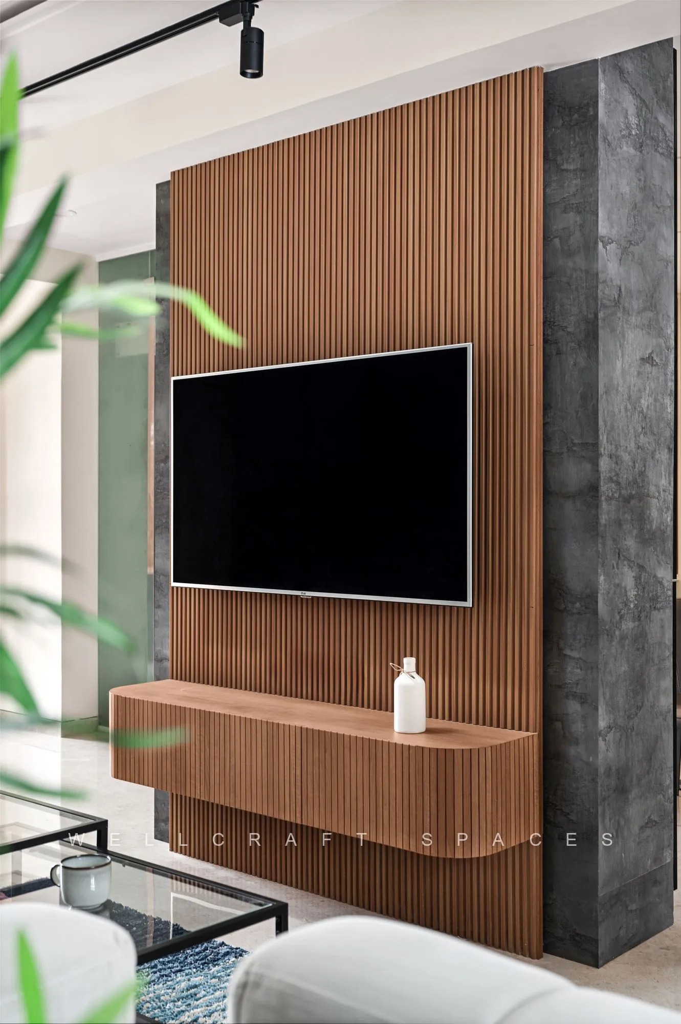 Media wall of fluted walnut panelling flanked by grey concrete-finish slabs, with a wall-mounted television, a floating ledge and a white curved sofa.