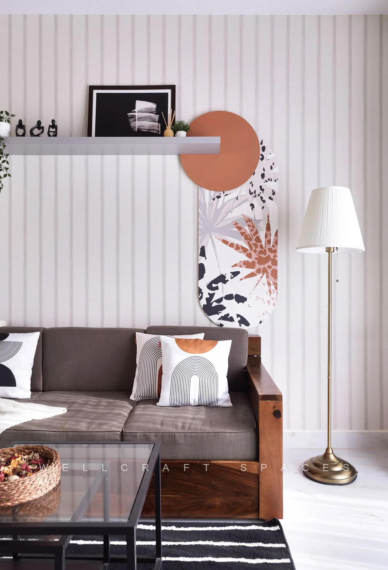 Seating detail with printed cushions, a floating grey ledge holding a framed print and a terracotta disc, and a slim brass floor lamp beside the sofa.