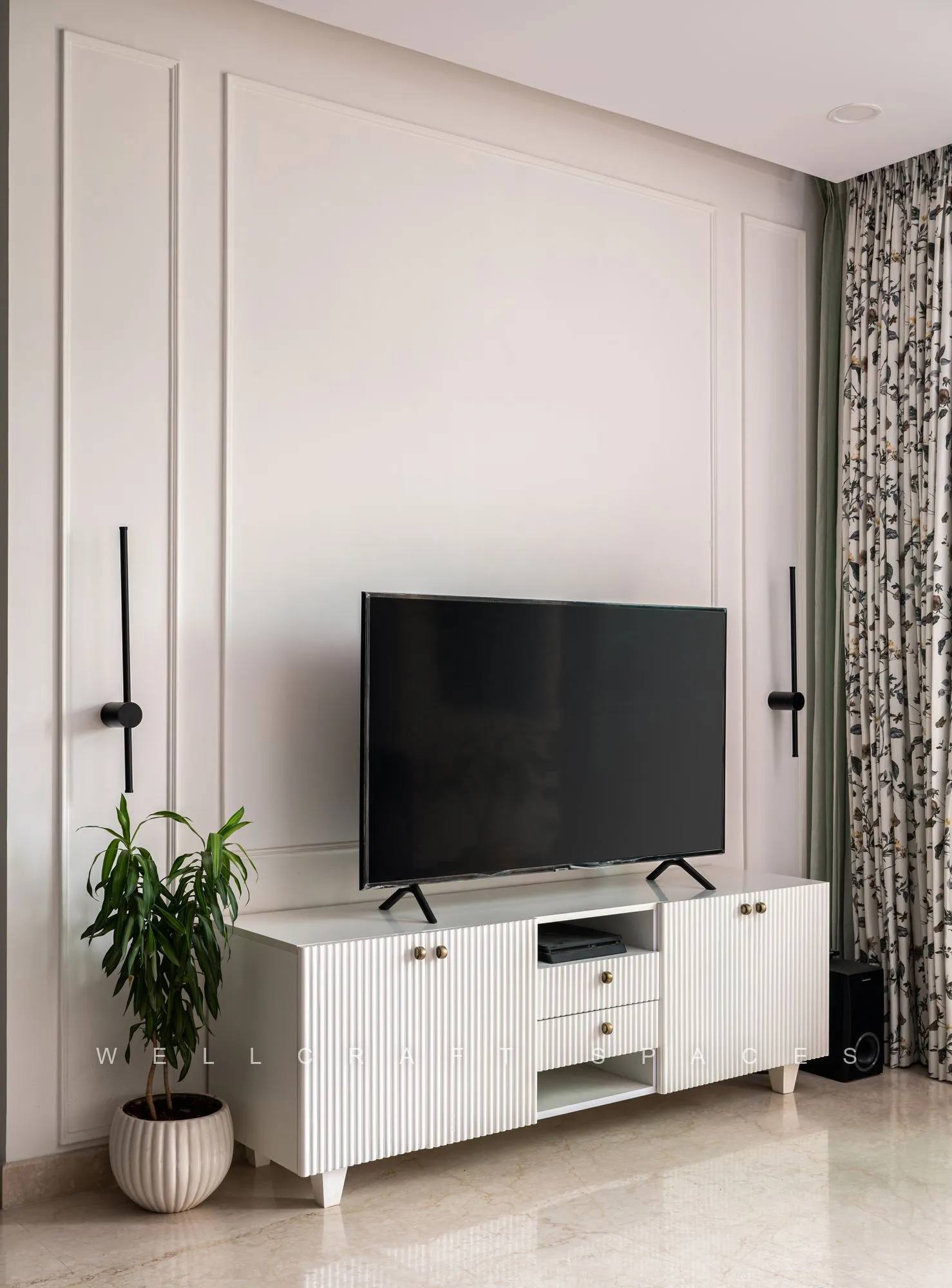 Media console in fluted white with brass handles beneath a wall-mounted television, flanked by black wall sconces and botanical-print curtains.