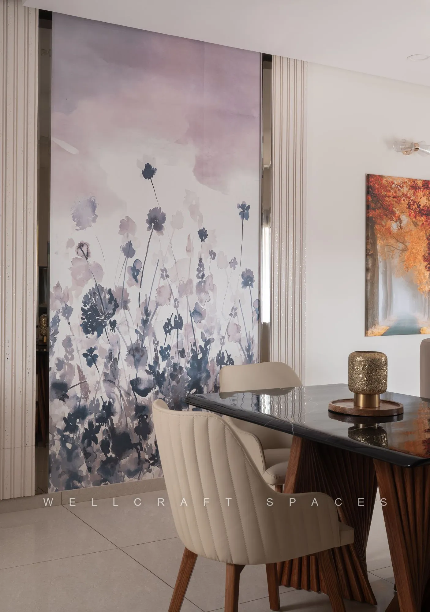 Dining corner set against a large blue-and-blush floral mural, with a dark glass-top table, fluted upholstered chairs and a brass table lamp.