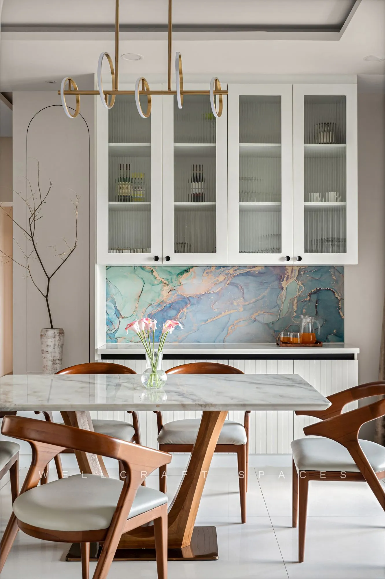 Dining table with a pale marble top and cane-backed wooden chairs under brass pendant arms, framed by glass-fronted white crockery cabinets.