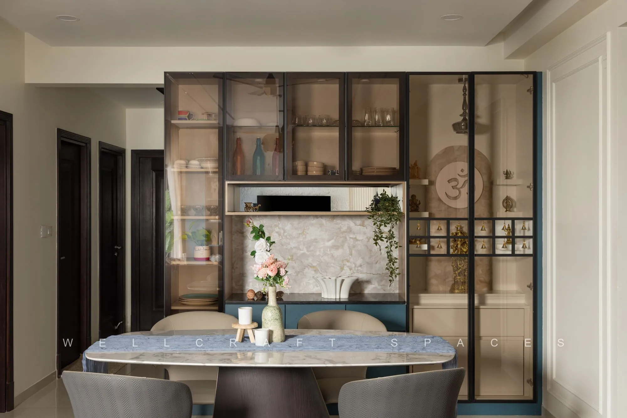 Round dining table with grey chairs in front of a glass-fronted crockery unit in teak and black metal, styled with pottery and flowers.