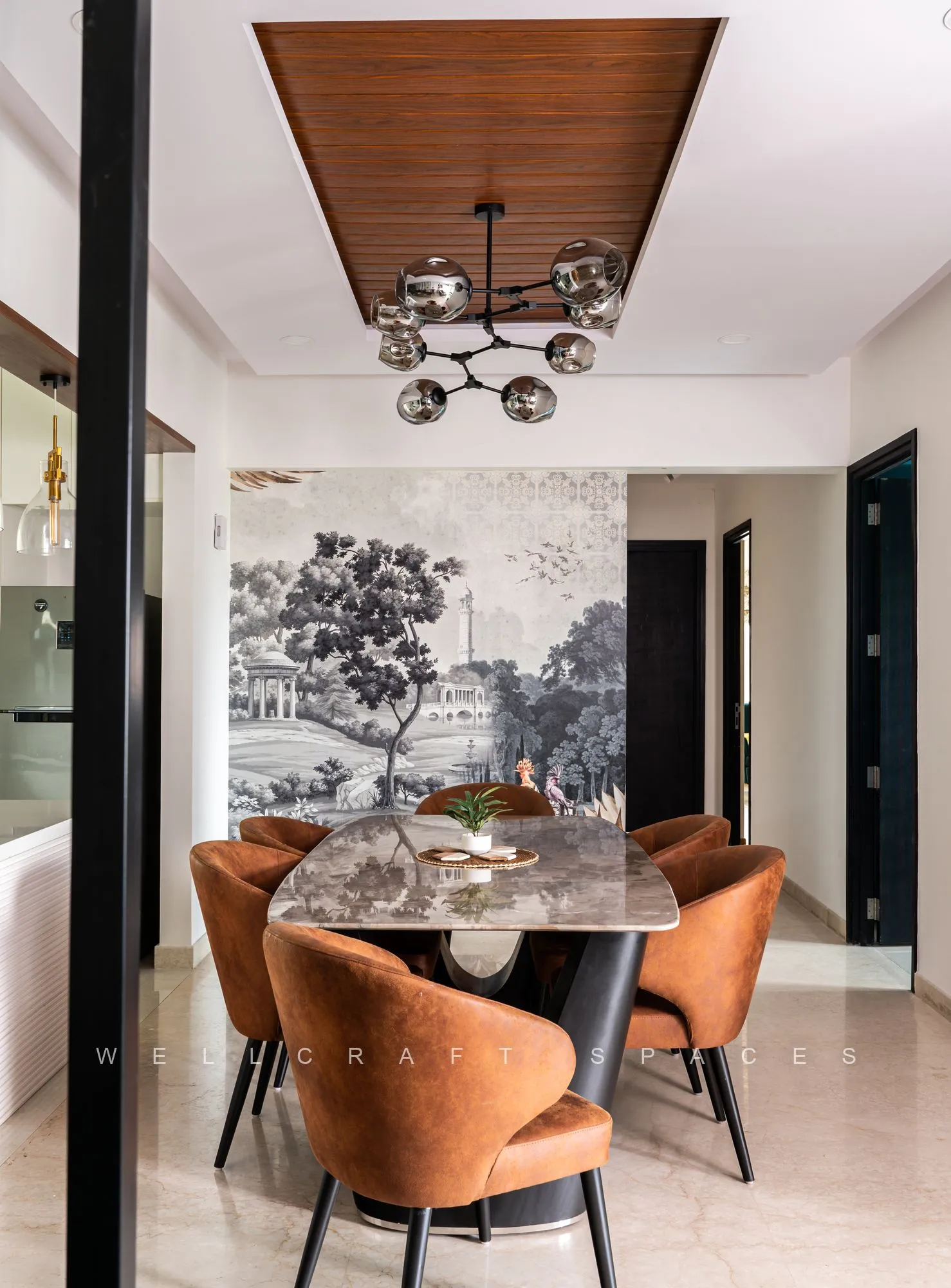 Round marble-top dining table with rust velvet chairs under a black sputnik chandelier, against a monochrome landscape mural and a timber-slatted ceiling.
