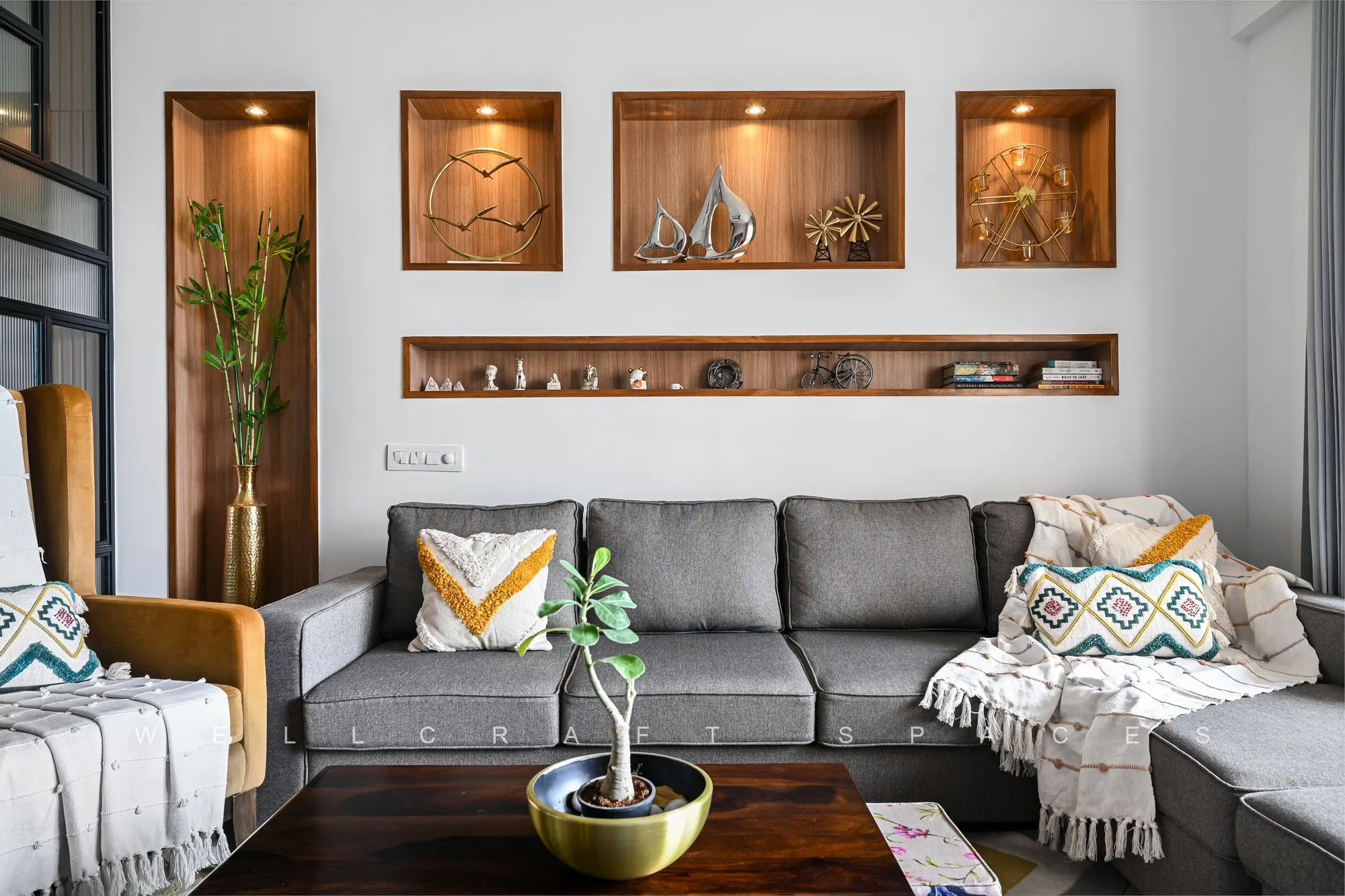 Living room feature wall with backlit walnut display niches holding brass and steel ornaments, a long curio shelf, and a grey sectional dressed with block-printed cushions.
