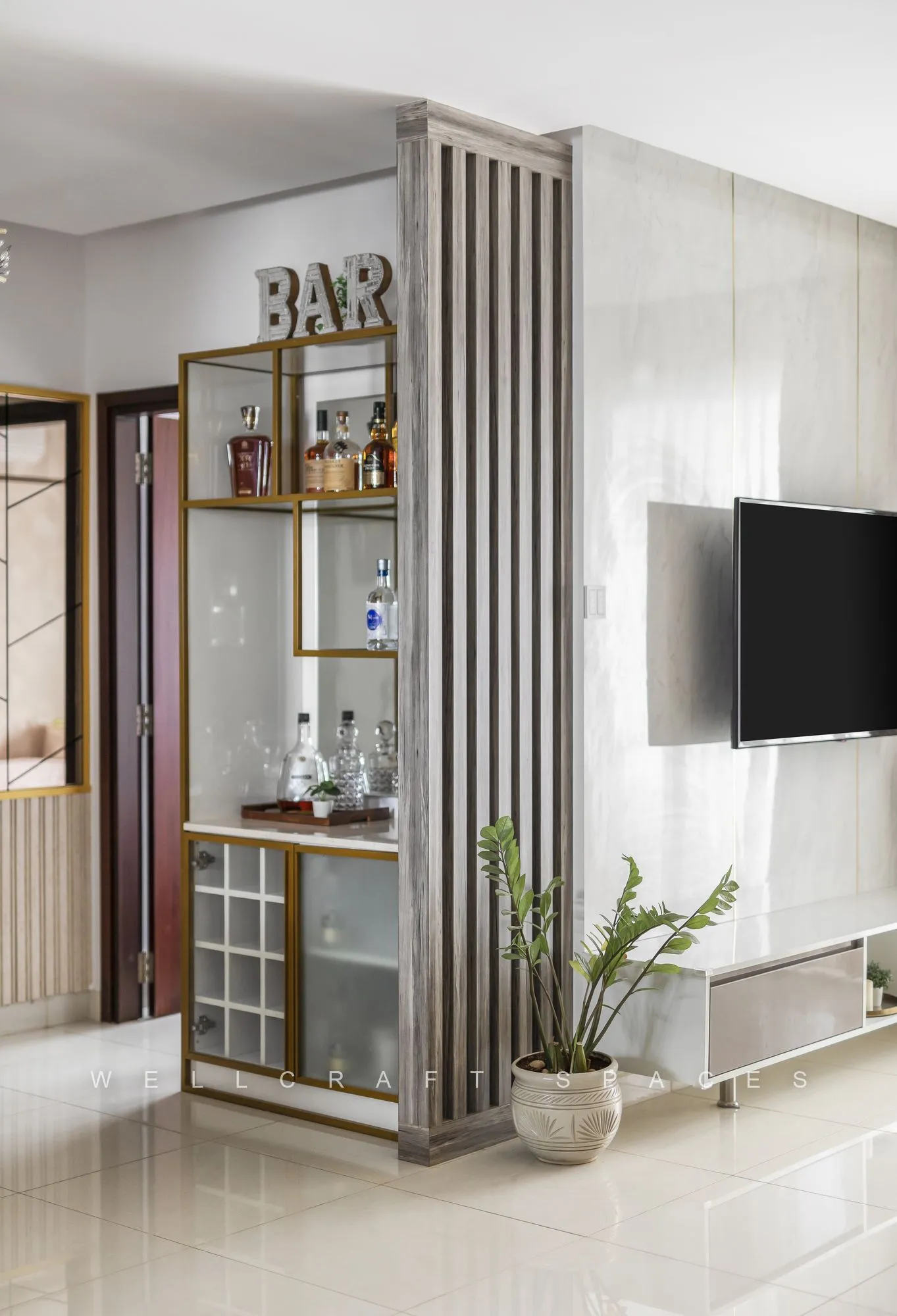 Mirror-backed home bar with brass-framed shelves and glassware, set beside a fluted column partition and the television wall.