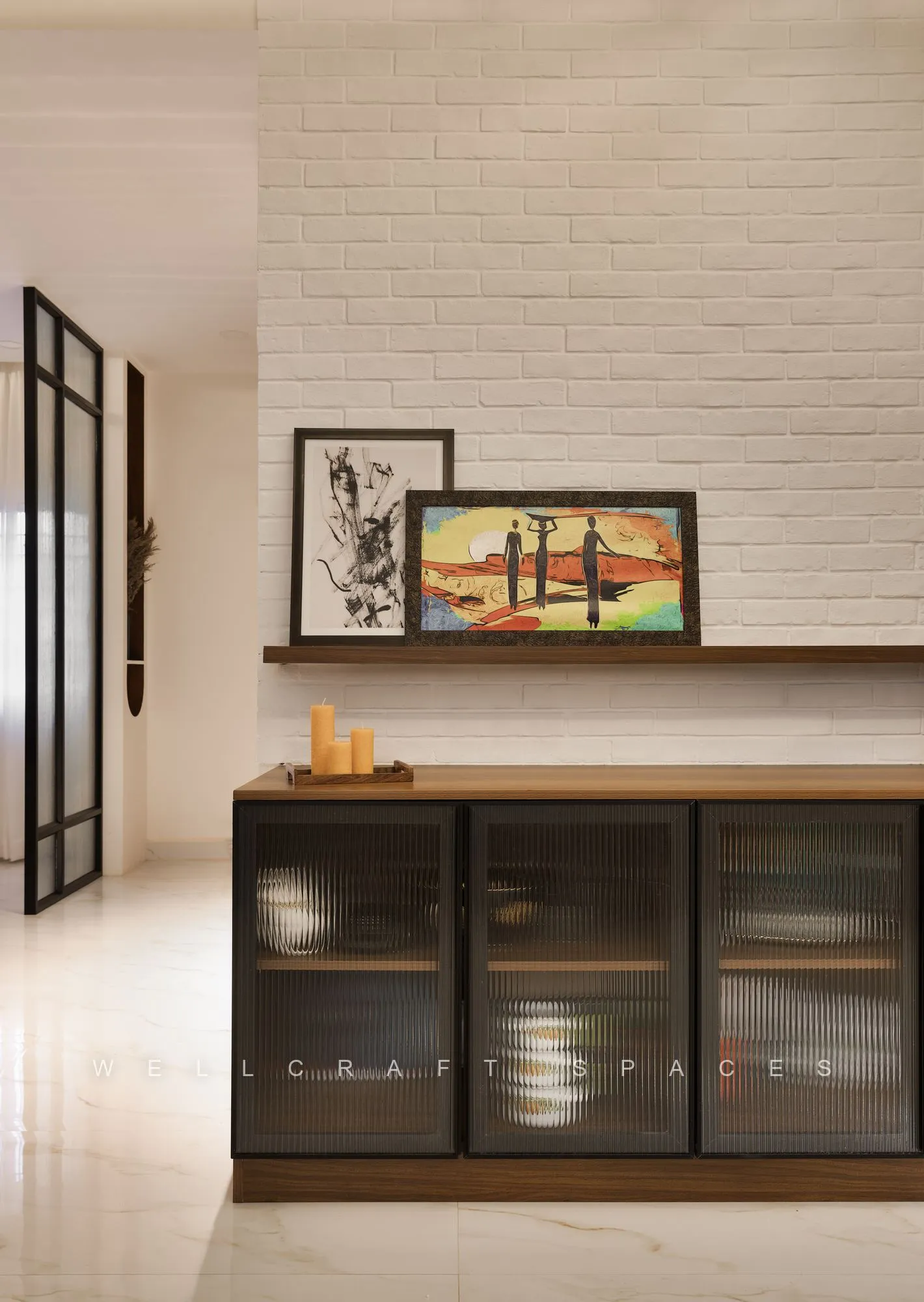 Dining sideboard in walnut with smoked fluted-glass shutters, a framed landscape painting and brass candlesticks against a textured white brick wall.