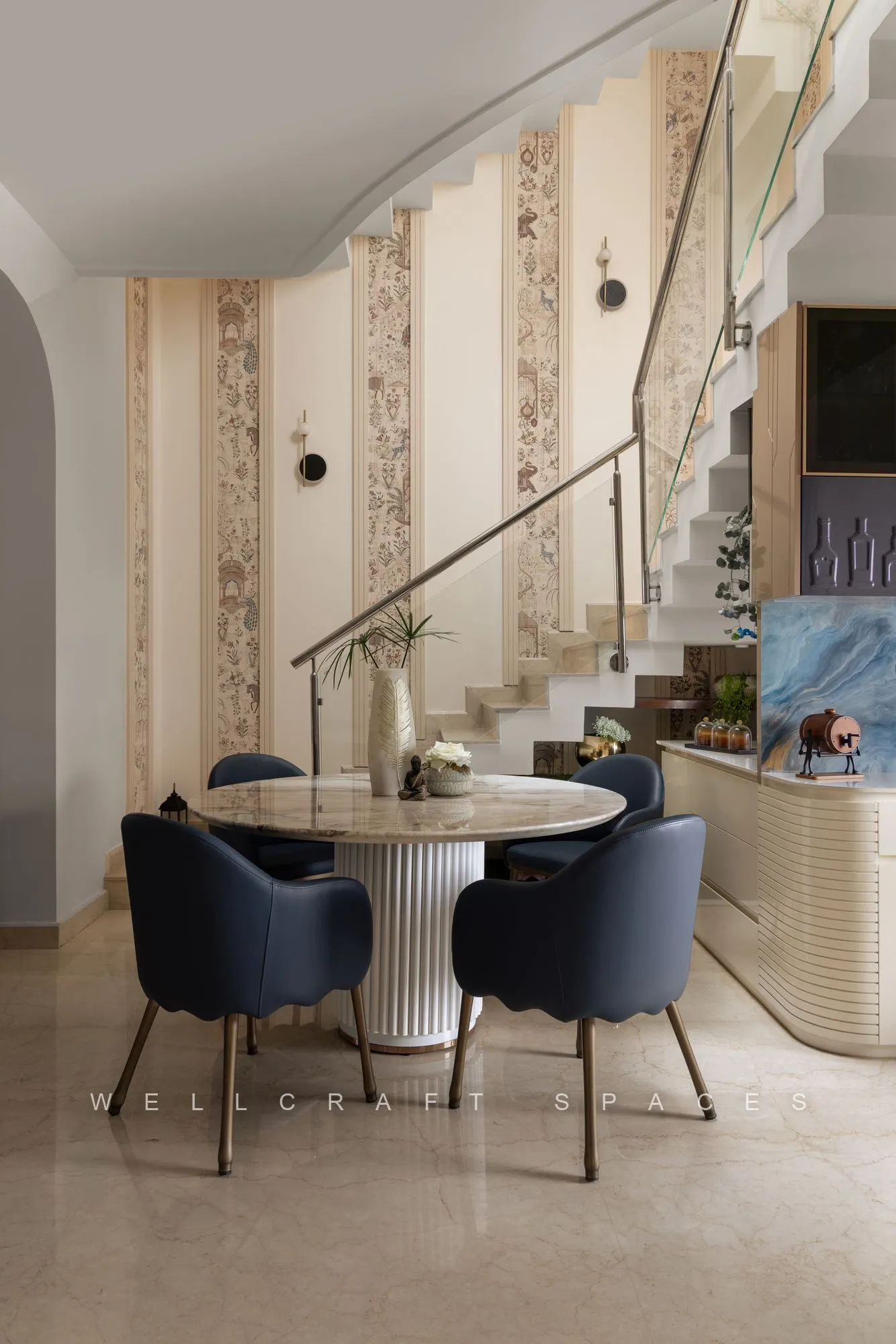 Round dining table with navy velvet chairs tucked beneath the staircase, against striped wallpaper and a slim brass railing.