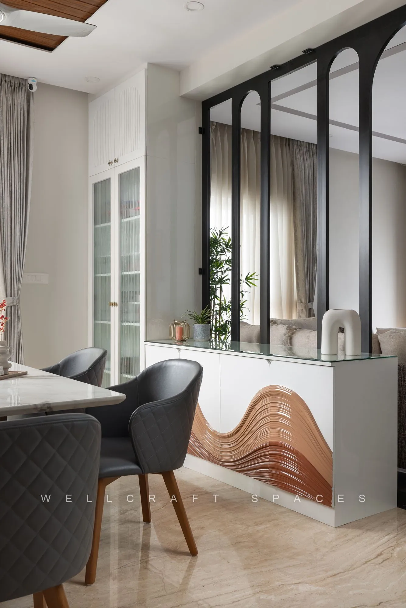 Dining chairs in grey quilted upholstery beside a sculptural wave-fronted timber console, framed by black arched glazing and trailing greenery.