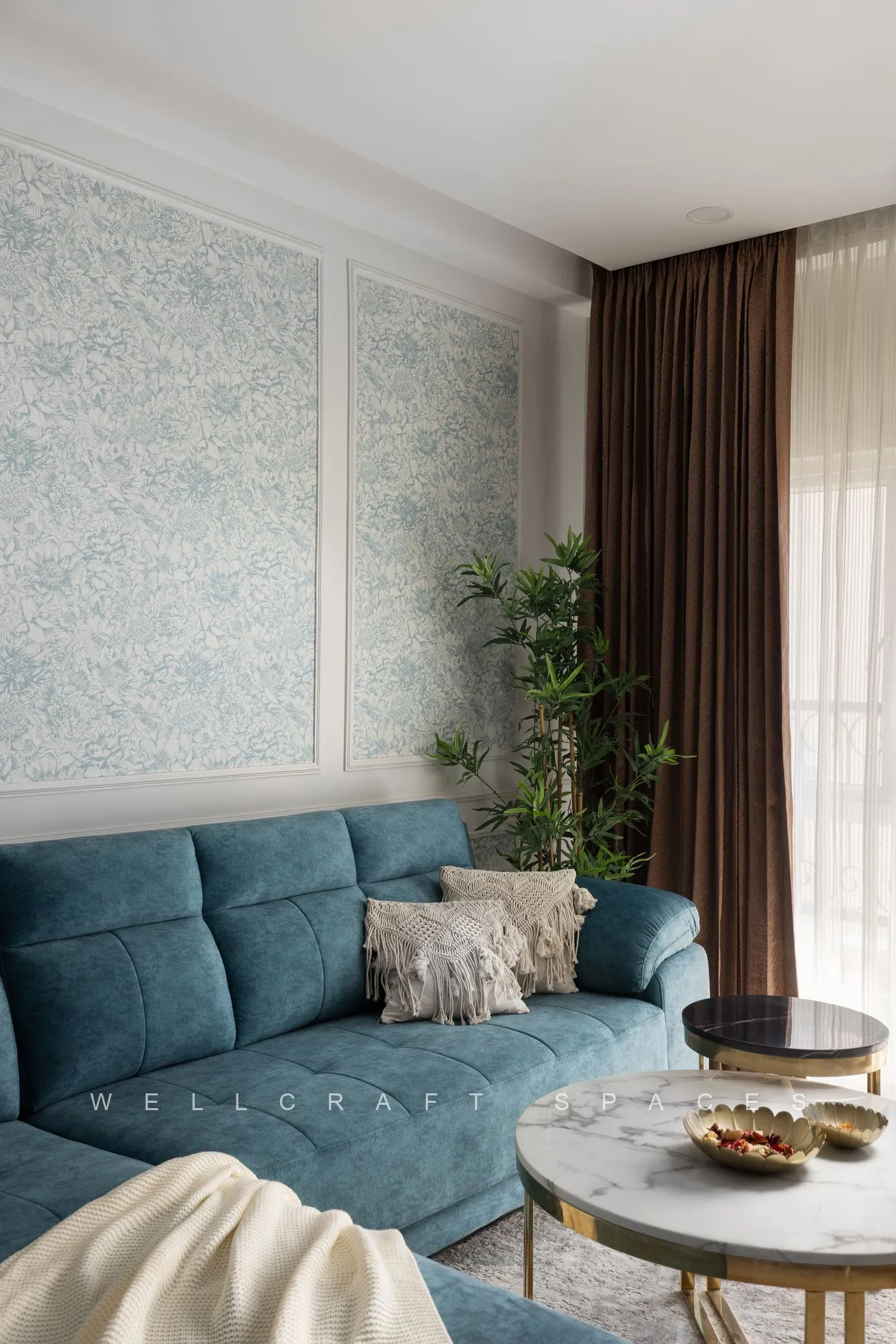 Teal three-seater sofa with a cream throw, a round marble coffee table and floor-length brown drapes beside a leafy plant.