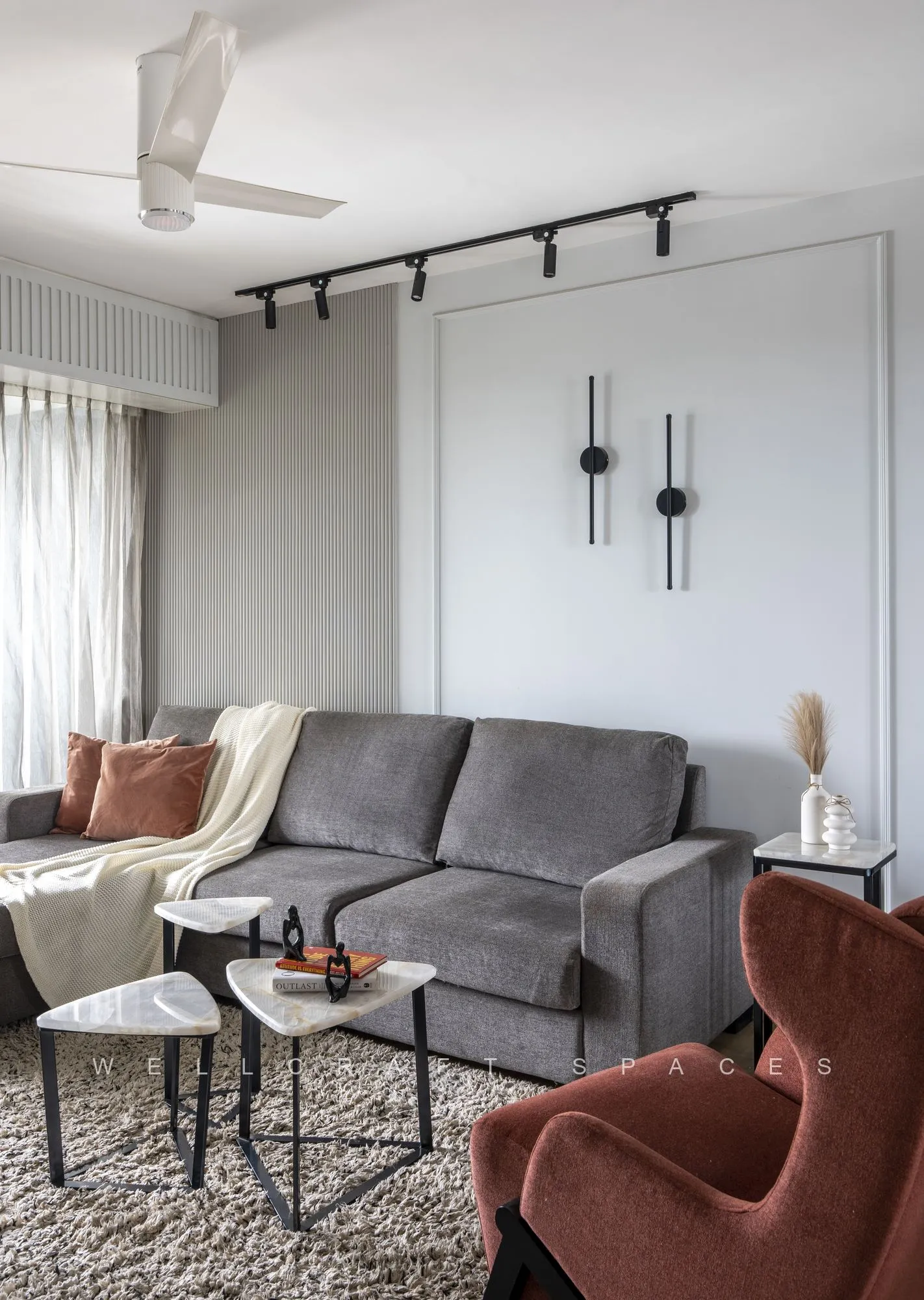 Grey three-seater with rust cushions flanked by terracotta velvet wing chairs, nested black metal side tables and slim black wall sconces on panelled walls.