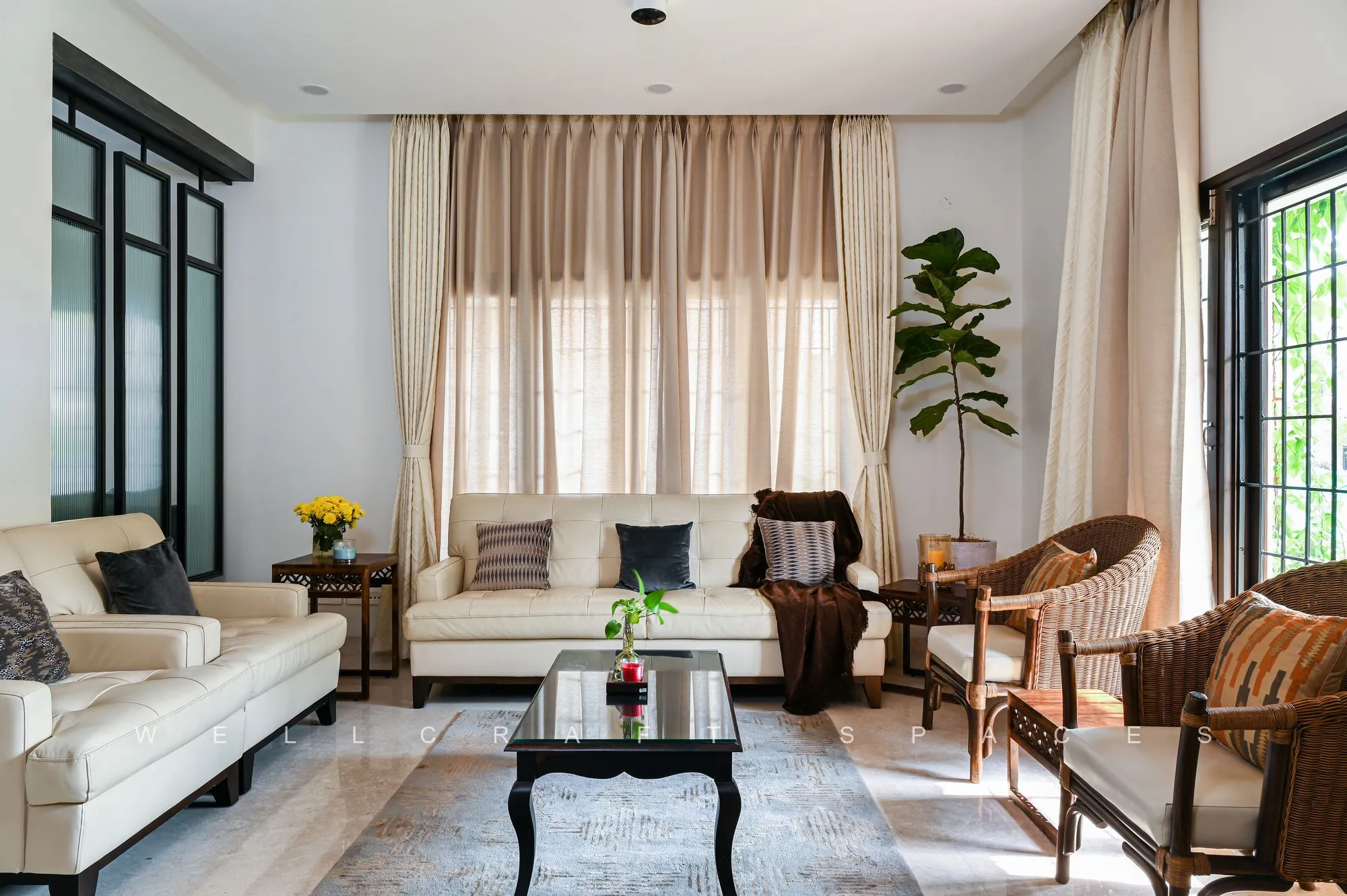 Bright living room with a cream sofa, cane-and-teak armchairs, a low black coffee table and a hand-knotted rug, lit by tall curtained windows.