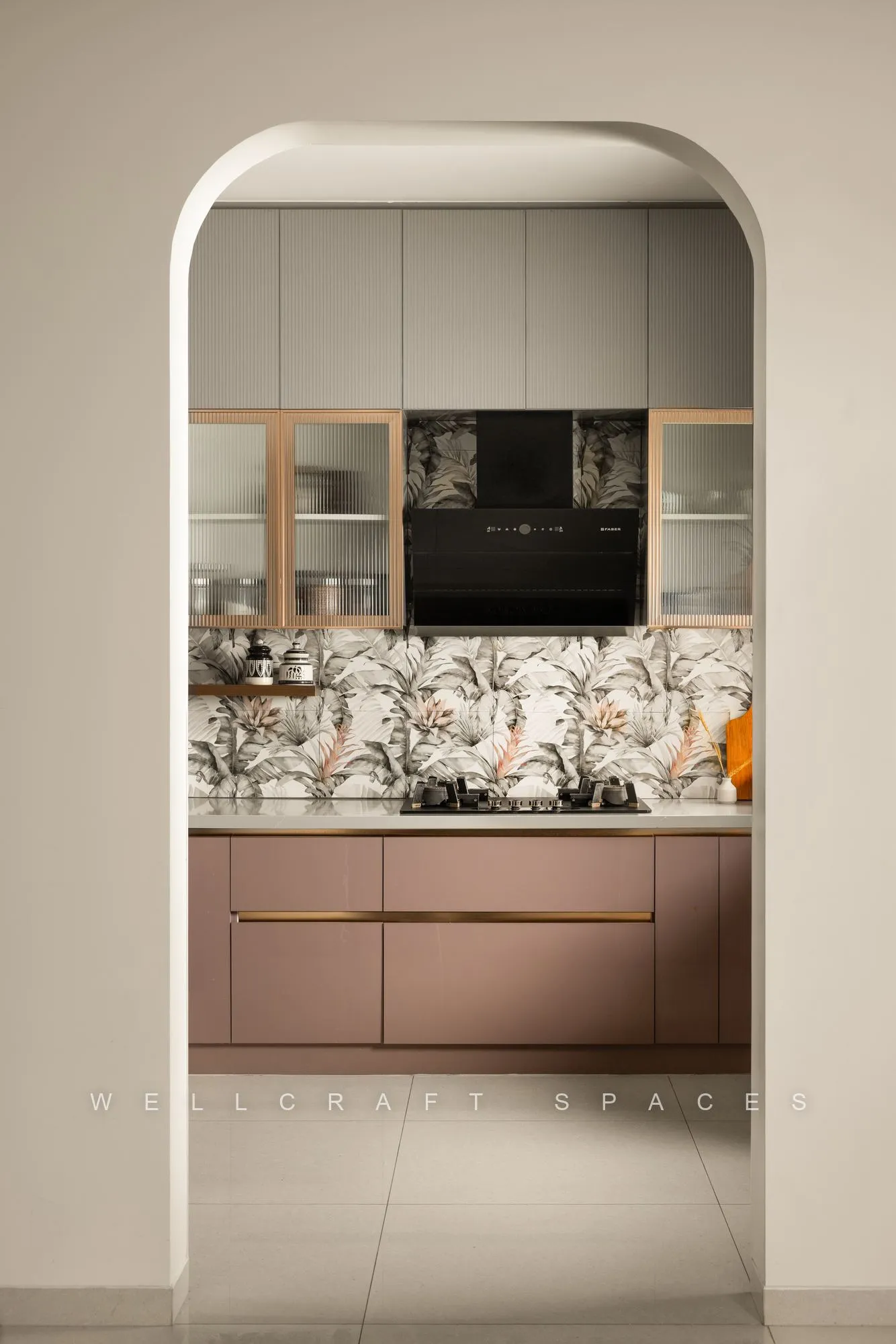 Kitchen seen through a tall plaster arch, showing dusty-rose base cabinets, a botanical tile backsplash and glass-fronted wall units.